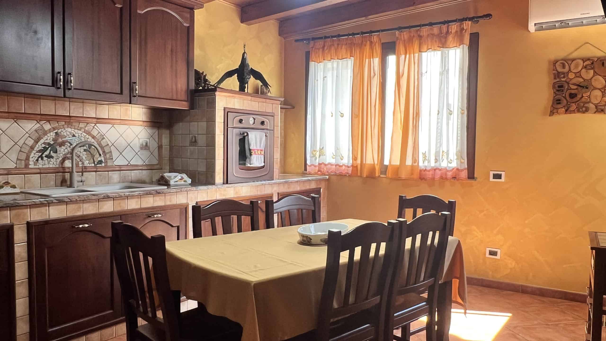 Interno di una cucina moderna con mobili in legno scuro, tavolo con tovaglia gialla e sedie nere, finestra con tende arancioni, pareti gialle e decorazioni.