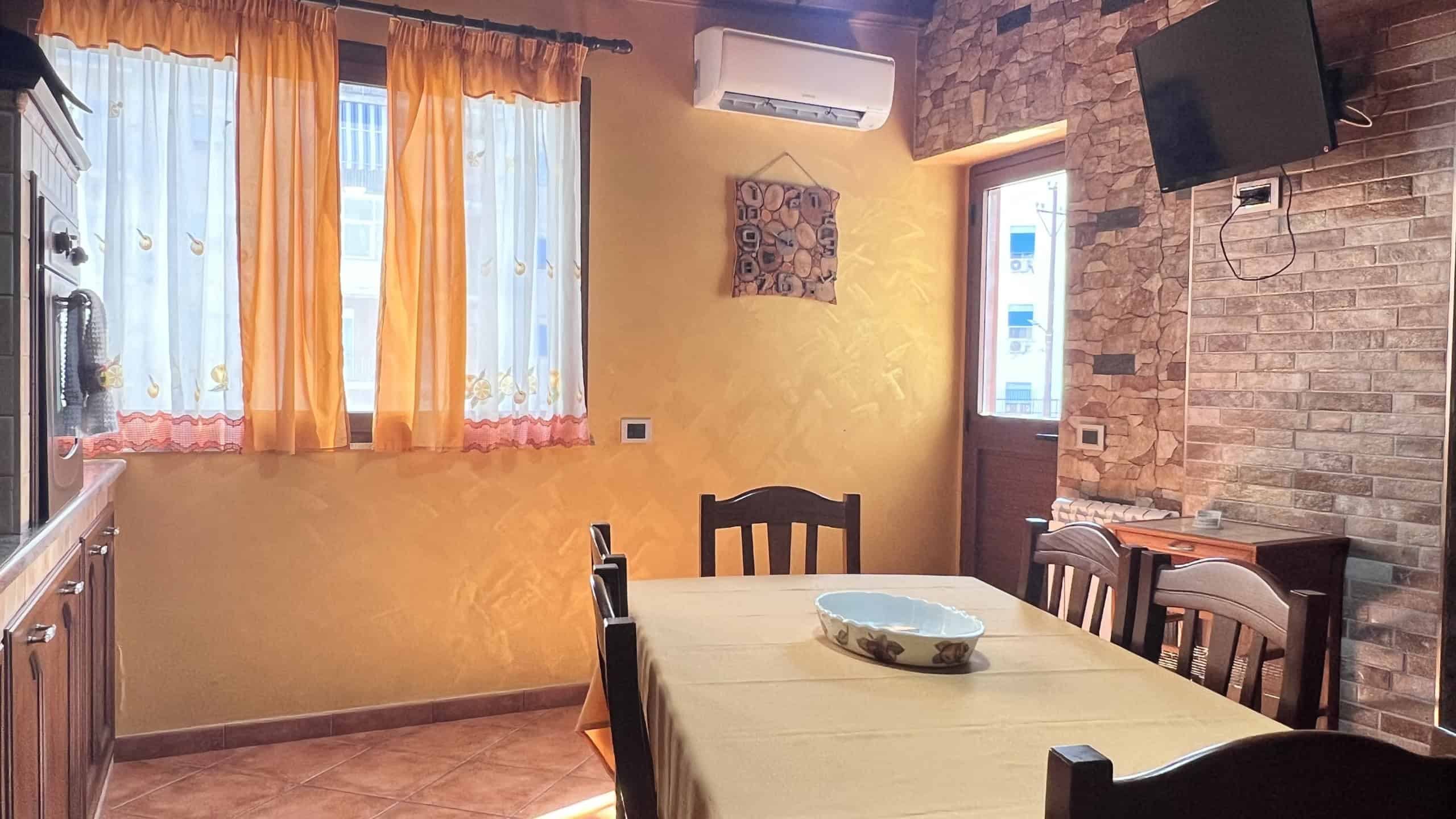 Interno di un appartamento con tavolo da pranzo, sedie, pareti gialle, finestra con tende arancioni e TV a schermo piatto.