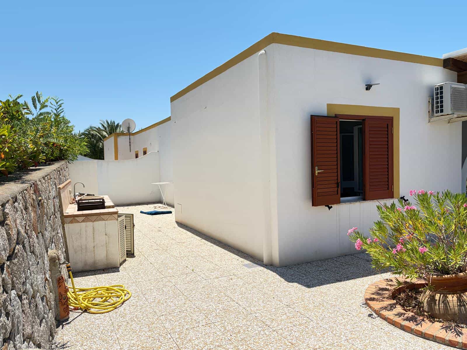 Esterno di una casa moderna con pareti bianche, finestra con persiana marrone e area esterna con piante e arredi da giardino.