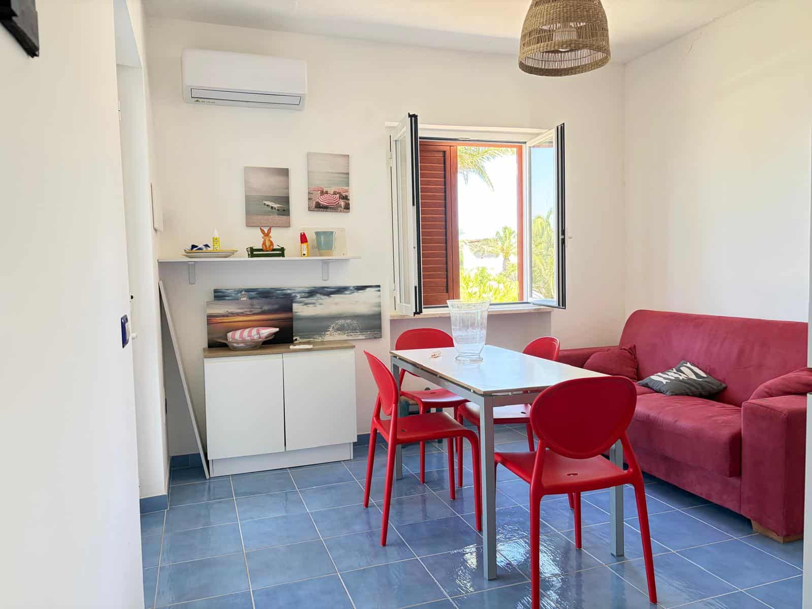 Interno di un appartamento con soggiorno, tavolo, sedie rosse, divano rosso e vista sul mare attraverso la finestra aperta.