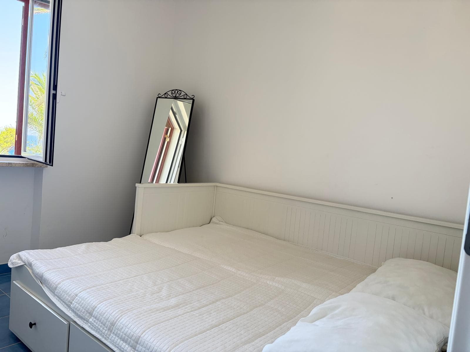 Camera da letto moderna con letto matrimoniale, finestra aperta e specchio decorativo, ambiente luminoso e accogliente.