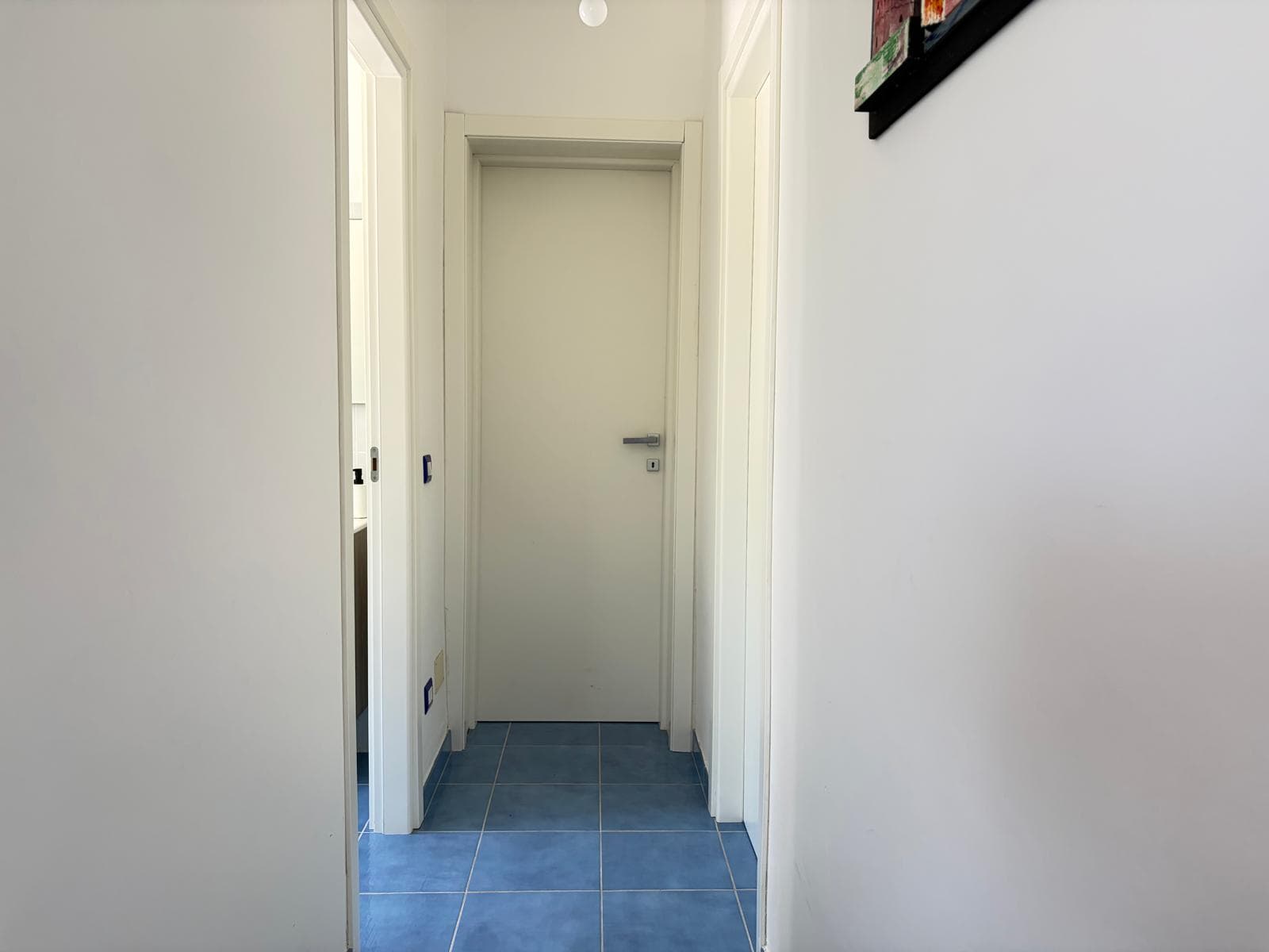 Corridor interno di una casa con pareti bianche e pavimento in piastrelle blu, porta chiusa alla fine.