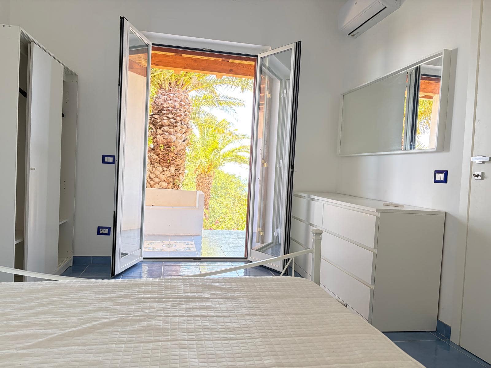 Camera da letto luminosa con porta finestra che si apre su un giardino con palme e vegetazione tropicale, arredamento minimalista bianco.