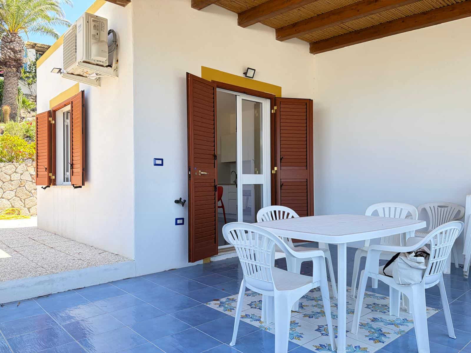 Spazio esterno con tavolo e sedie in plastica bianca, porta finestra con persiane in legno, aria condizionata, ambiente esterno di una casa al mare o in zona calda.