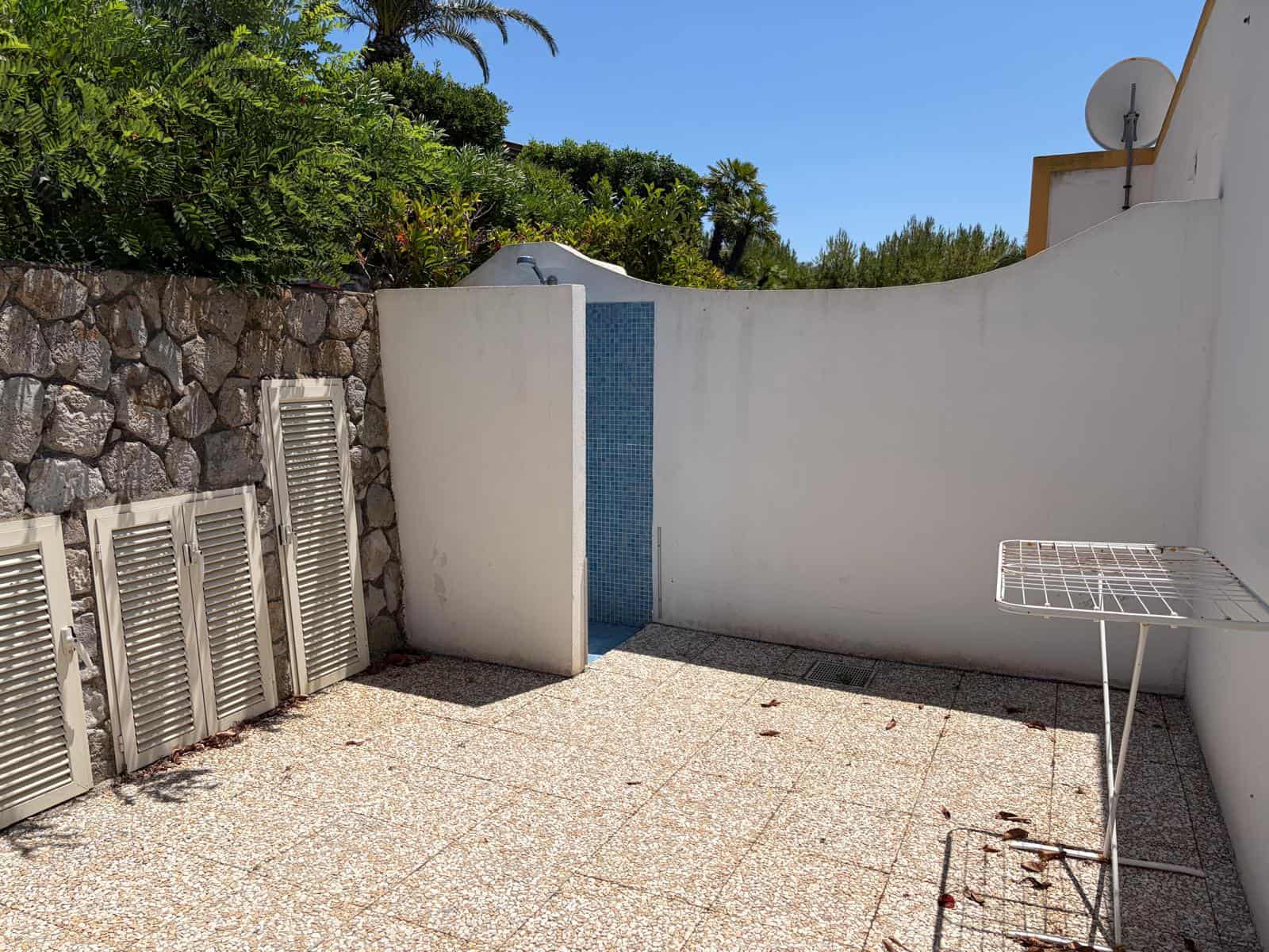 Spaziosa terrazza privata con pavimento in piastrelle, pareti bianche e vista su giardino e cielo azzurro, ideale per relax e convivialità all'aperto.