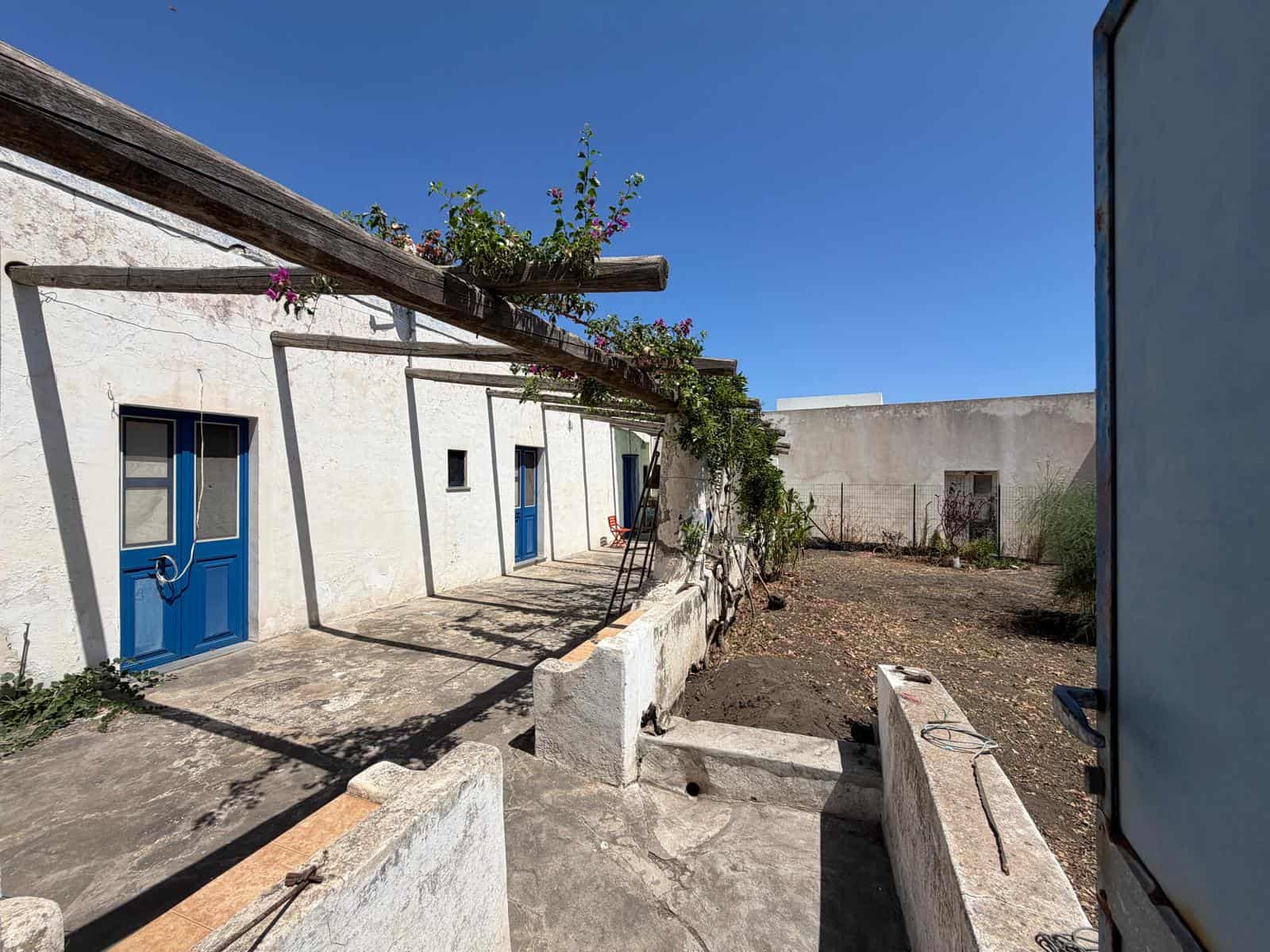Esterno di un cortile con pareti bianche, porte blu e piante rampicanti sotto un cielo azzurro.