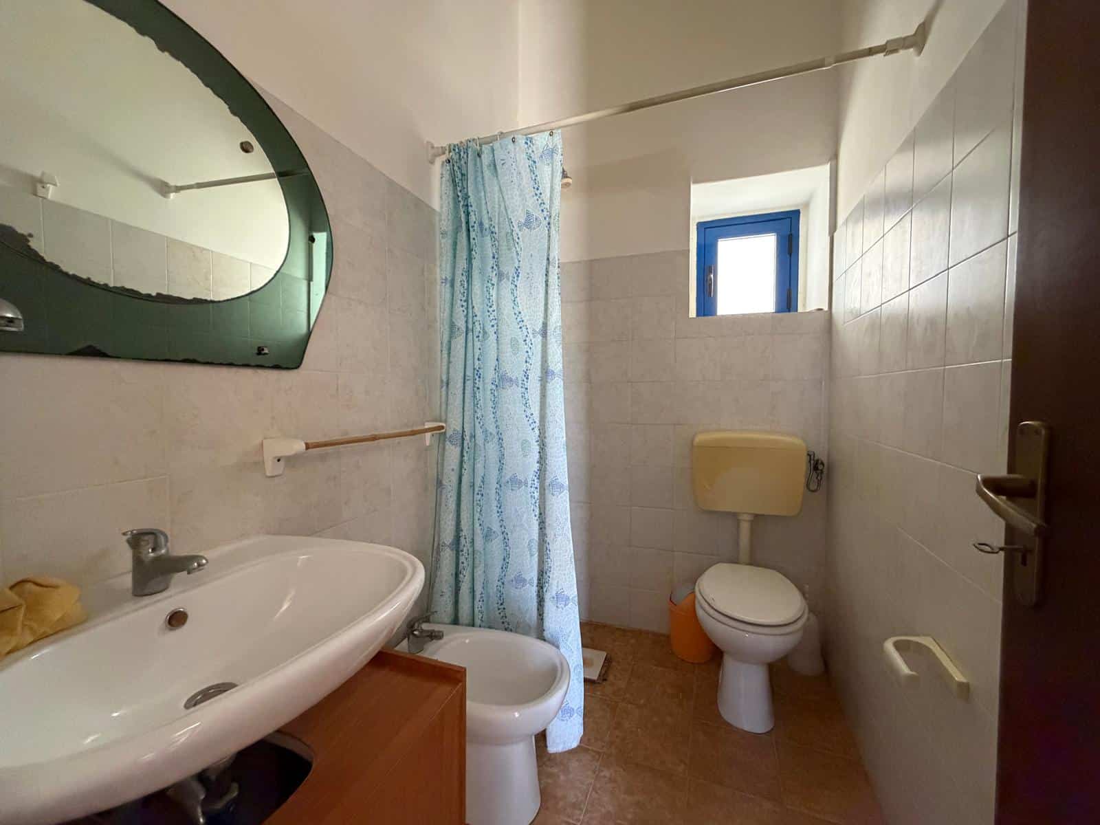 Bagno con lavabo, WC e doccia, finestra piccola, arredi semplici, ambiente funzionale.