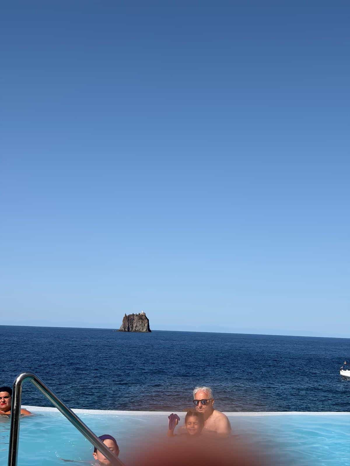 Persone che si godono una piscina con vista mare e un'isola rocciosa sullo sfondo, in una giornata soleggiata.