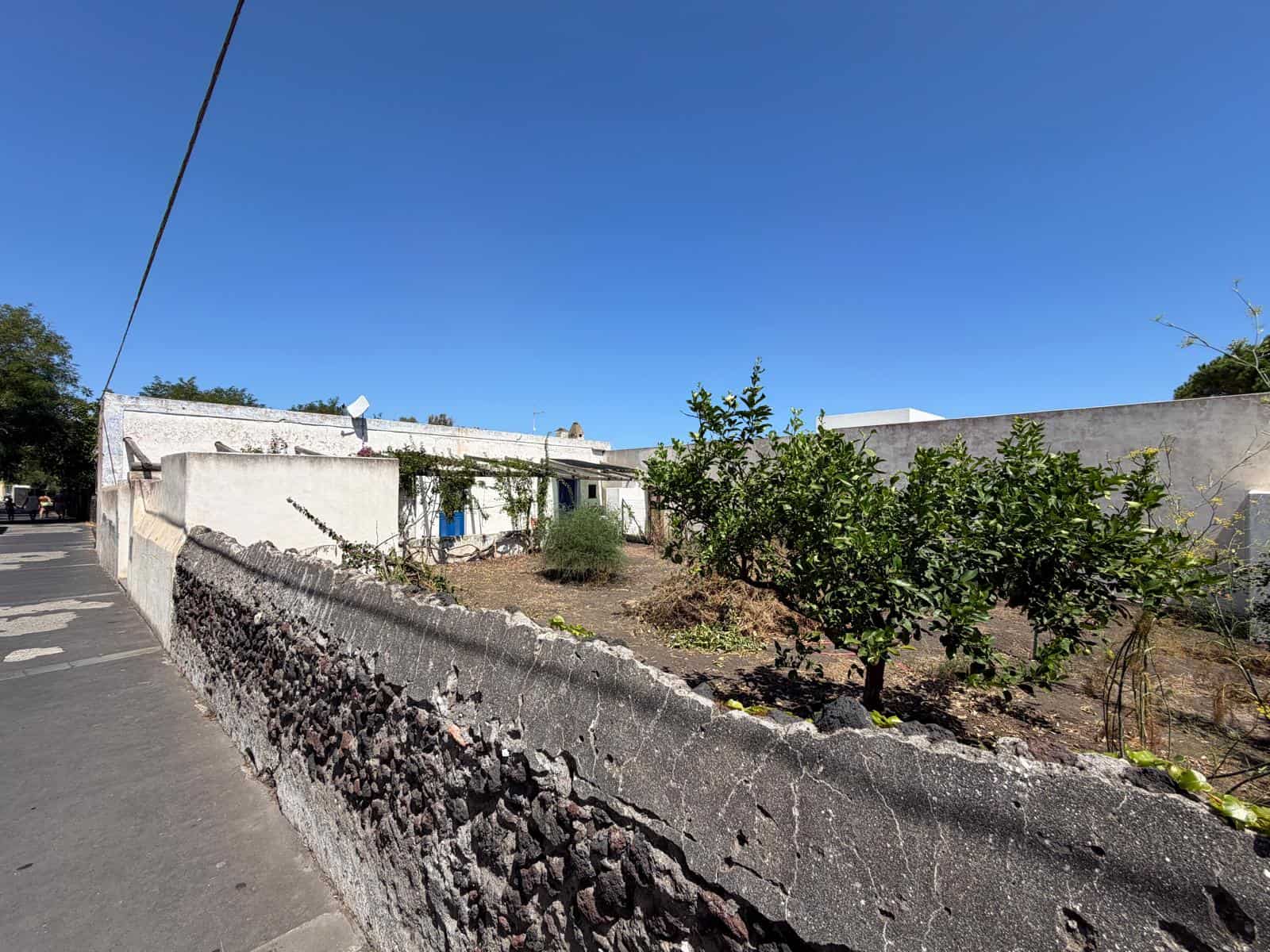 Giardino con alberi e muro di pietra sotto un cielo azzurro, vista di una zona residenziale in Italia.