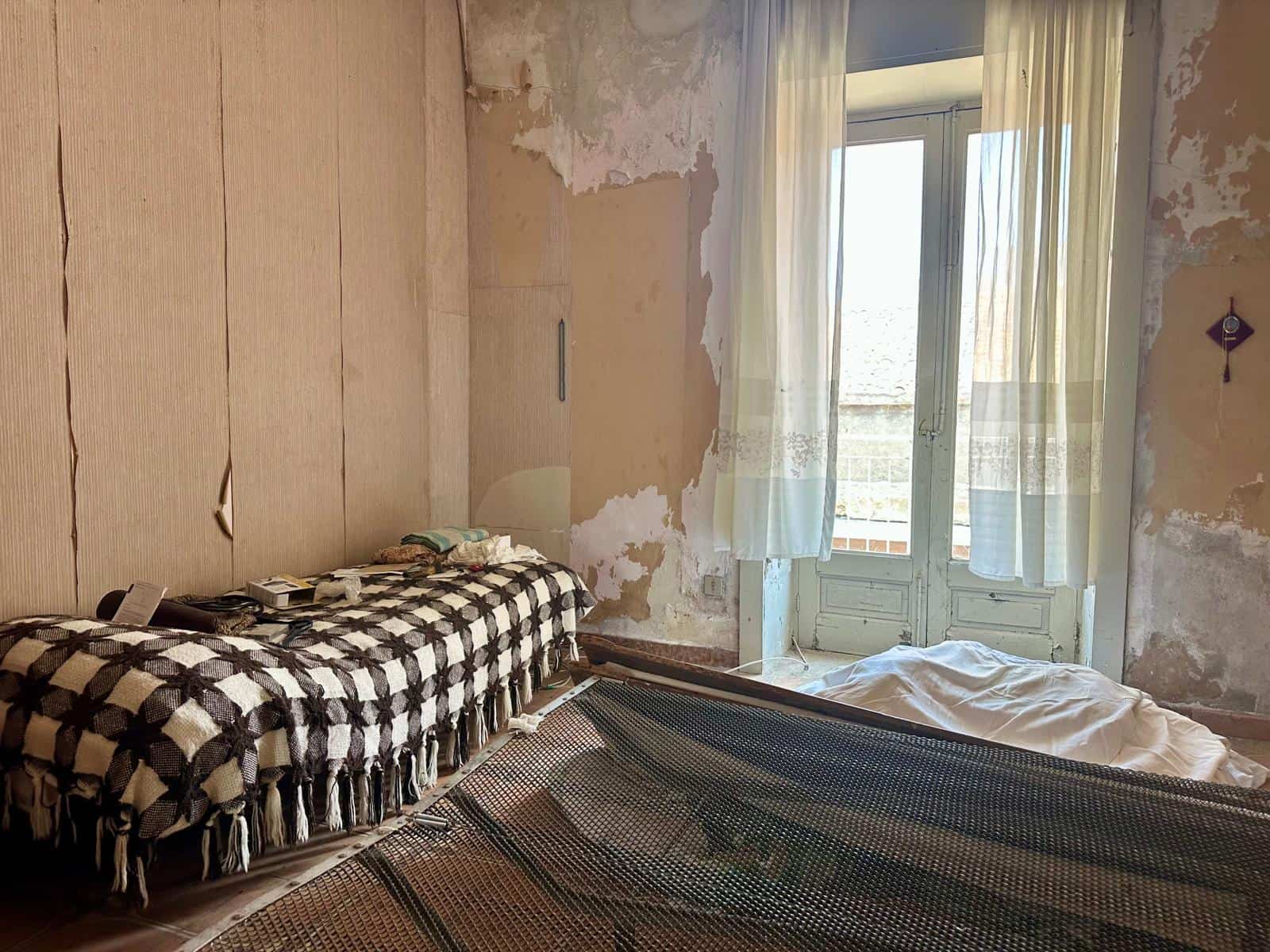 Interno di una casa in vendita con pareti danneggiate, letto e finestra con tende leggere.