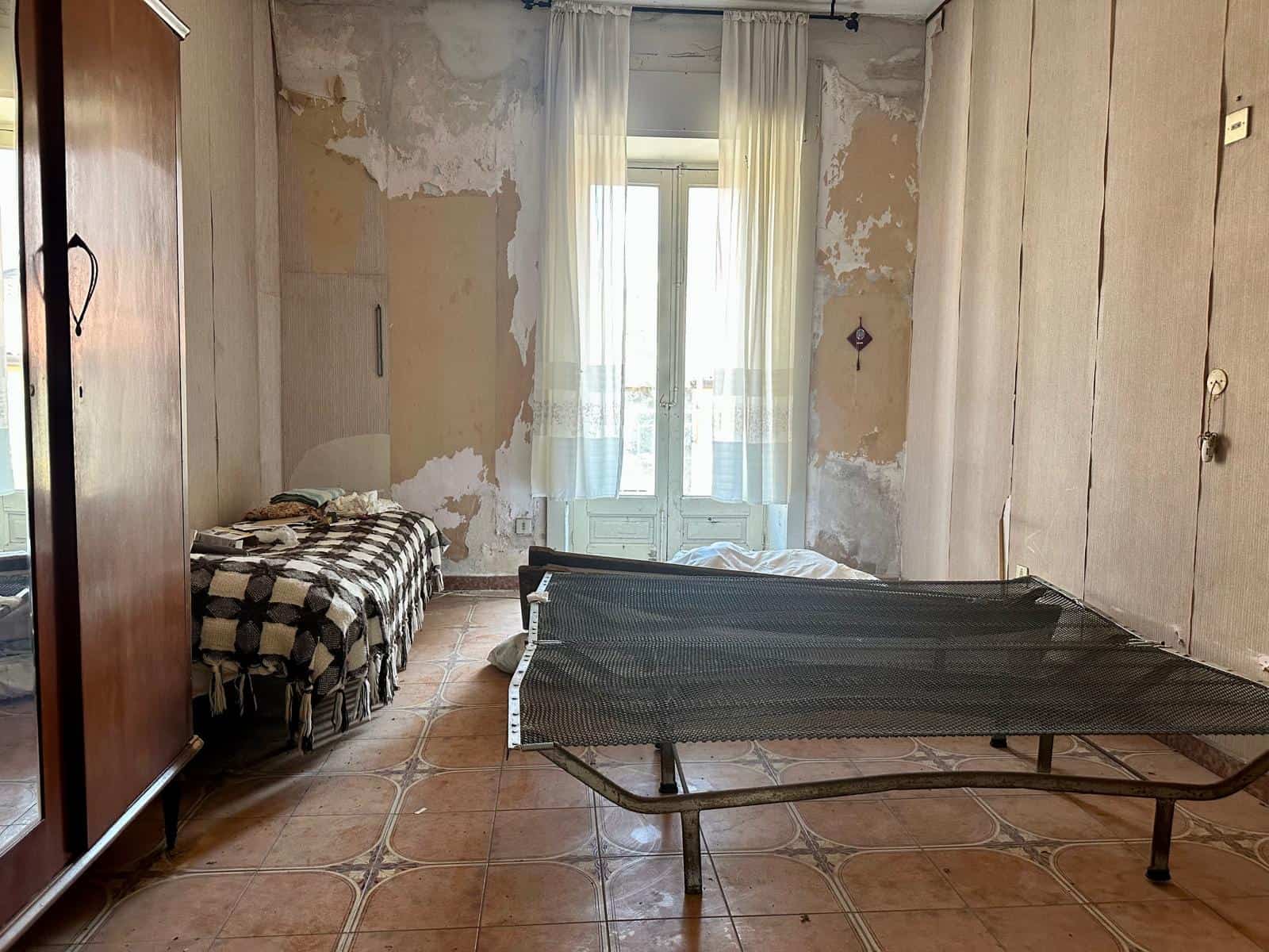 Interno di una casa indipendente in vendita con pareti danneggiate e un letto singolo, vista su una porta finestra con tende leggere.