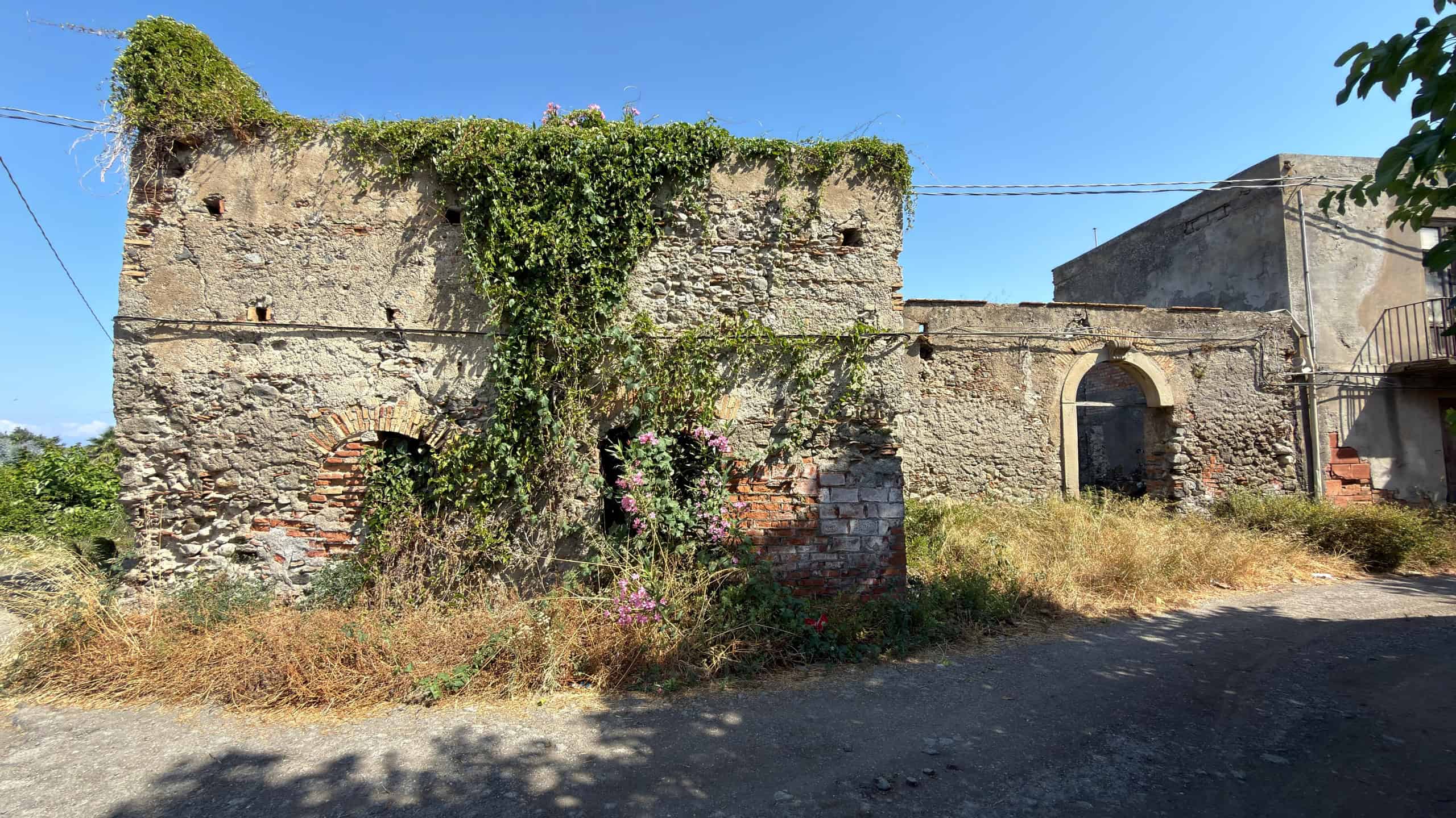 Vecchia struttura in pietra con vegetazione e piante rampicanti, situata in un'area rurale soleggiata.