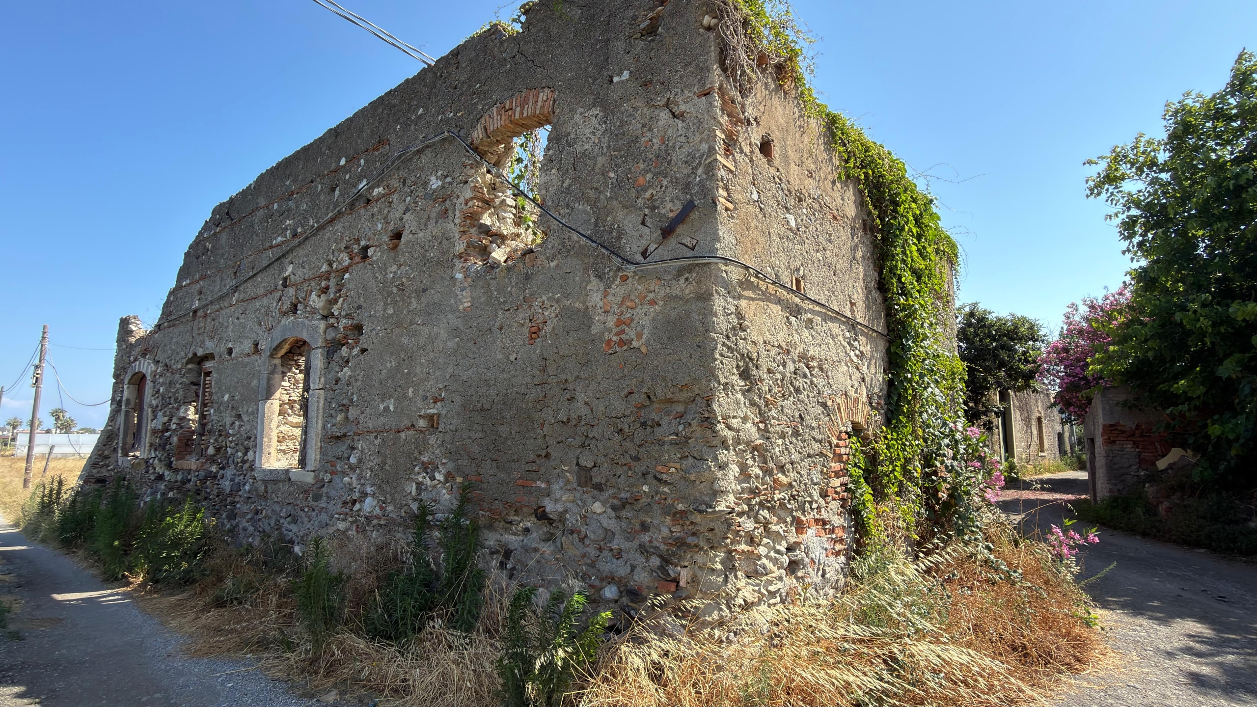 Vecchia casa in rovina con muri di pietra e finestre senza vetri, circondata da vegetazione e strada rurale in Italia.