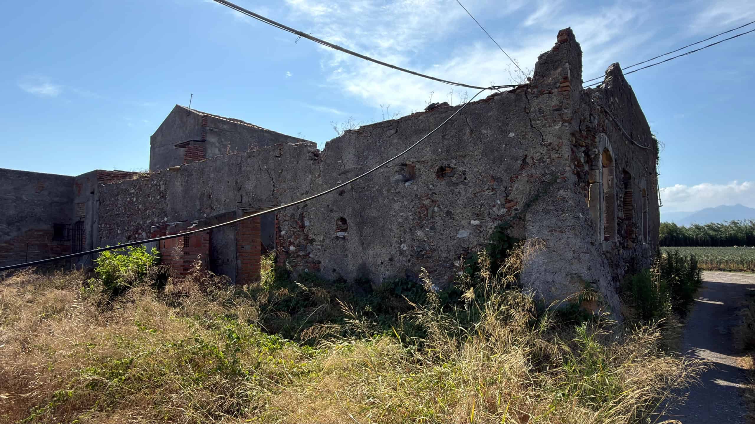 Vecchia casa abbandonata in campagna con muri in pietra e tetto in rovina, immersa in un paesaggio rurale soleggiato.