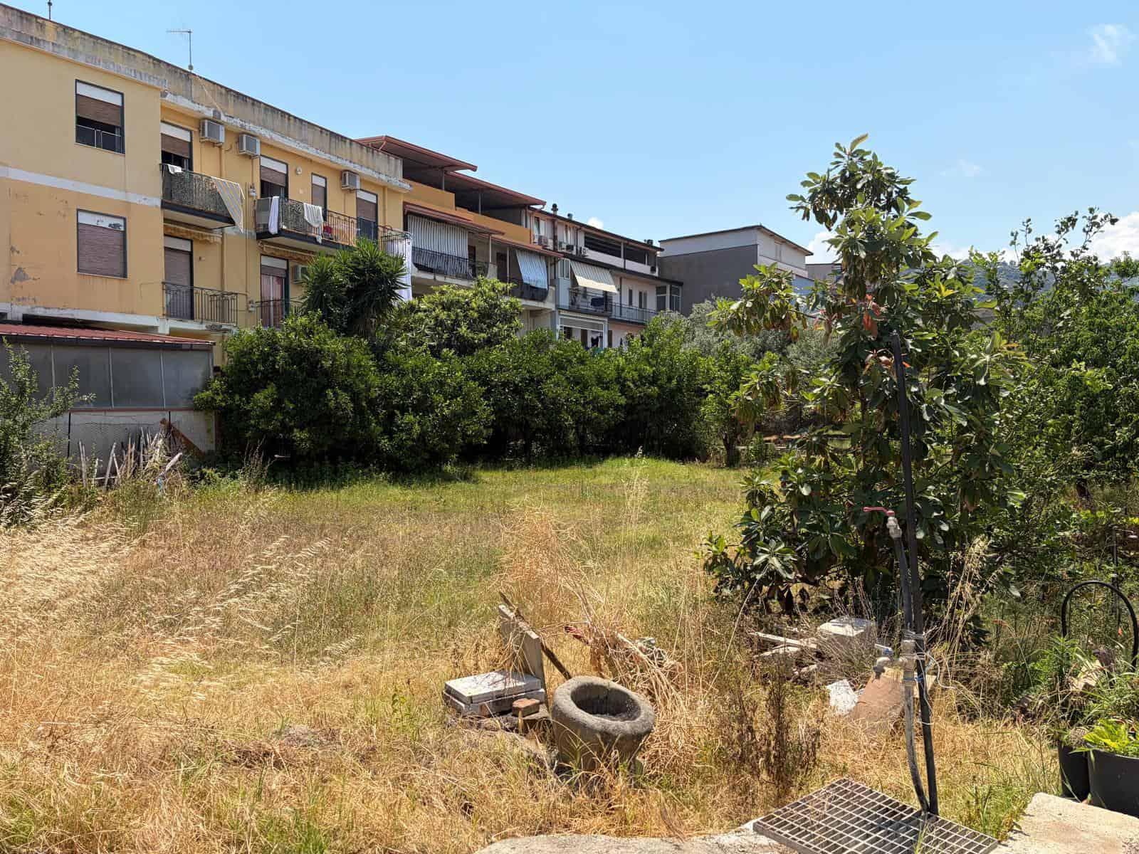 Terreno edificabile in vendita a Oliveri, con edifici residenziali sullo sfondo e vegetazione nel primo piano.
