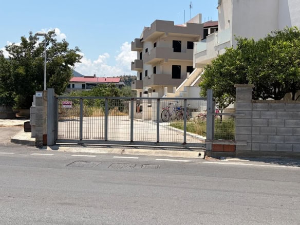 Ingresso di un terreno edificabile con cancello in metallo e strada asfaltata, vicino a edifici residenziali e vegetazione.
