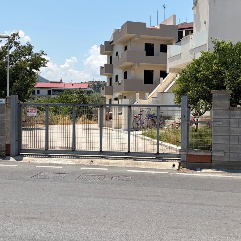 Ingresso di un terreno edificabile con cancello in metallo e strada asfaltata, vicino a edifici residenziali e vegetazione.