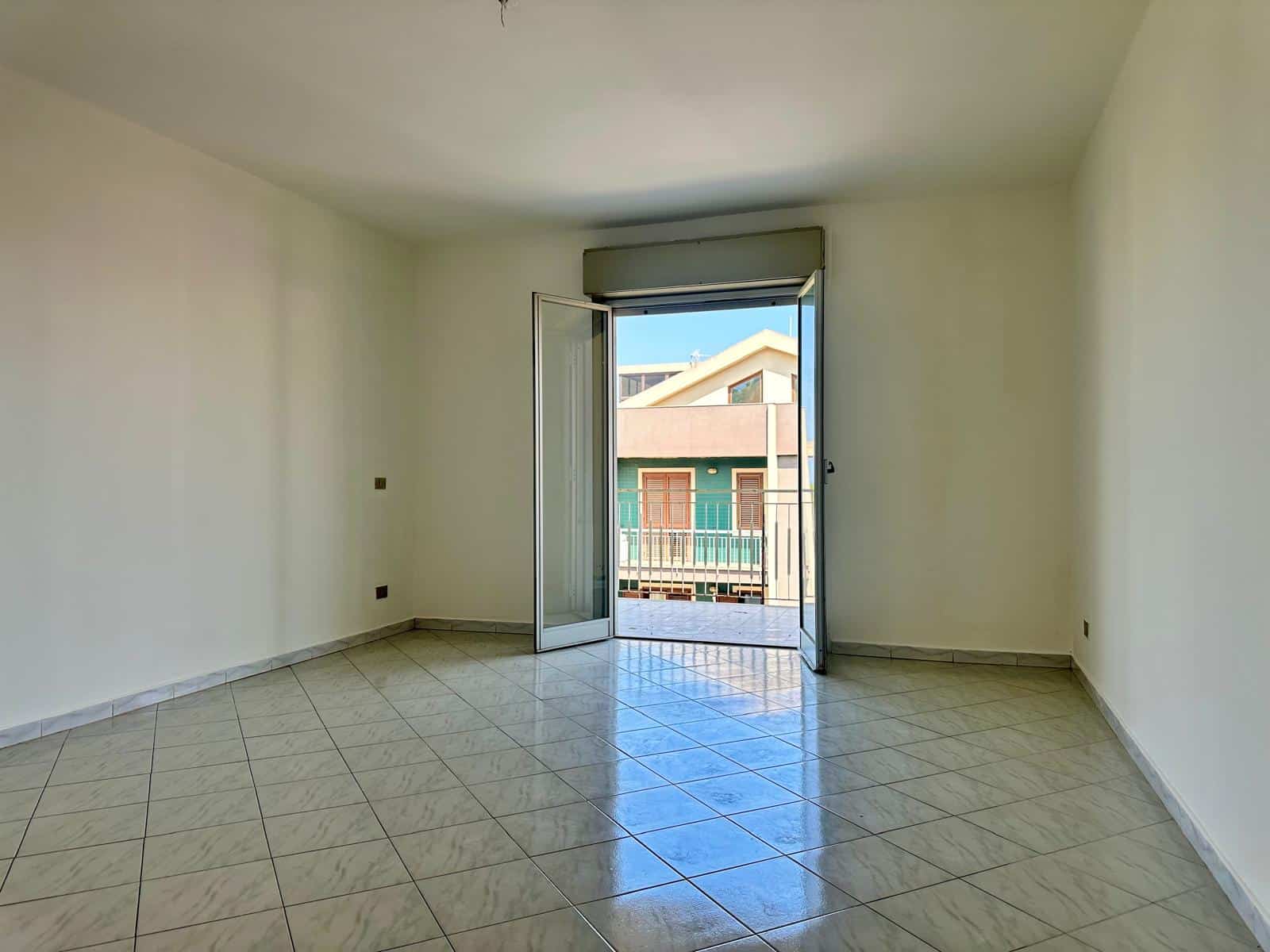 Stanza luminosa con porta finestra che dà su un balcone, pavimento in piastrelle e pareti bianche.