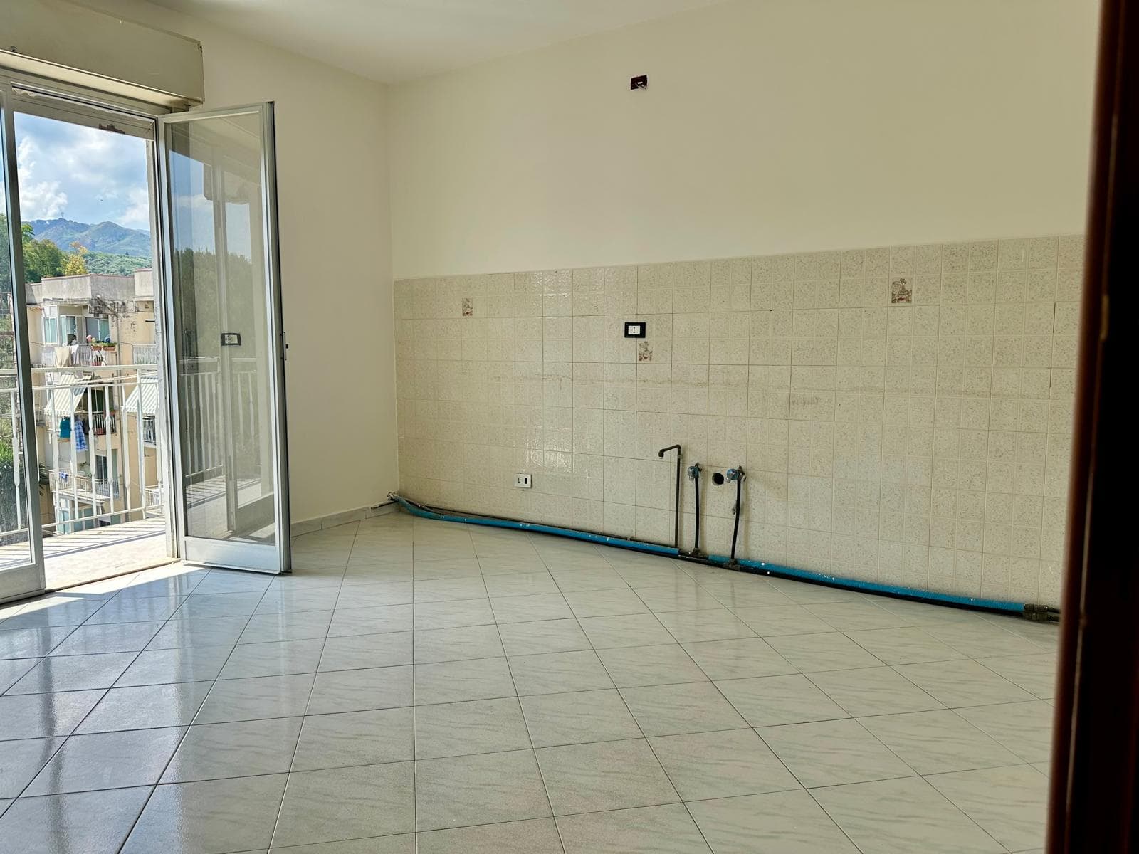 Interno di un appartamento in vendita con pareti bianche, pavimento in piastrelle e balcone con vista sulla città e le montagne.