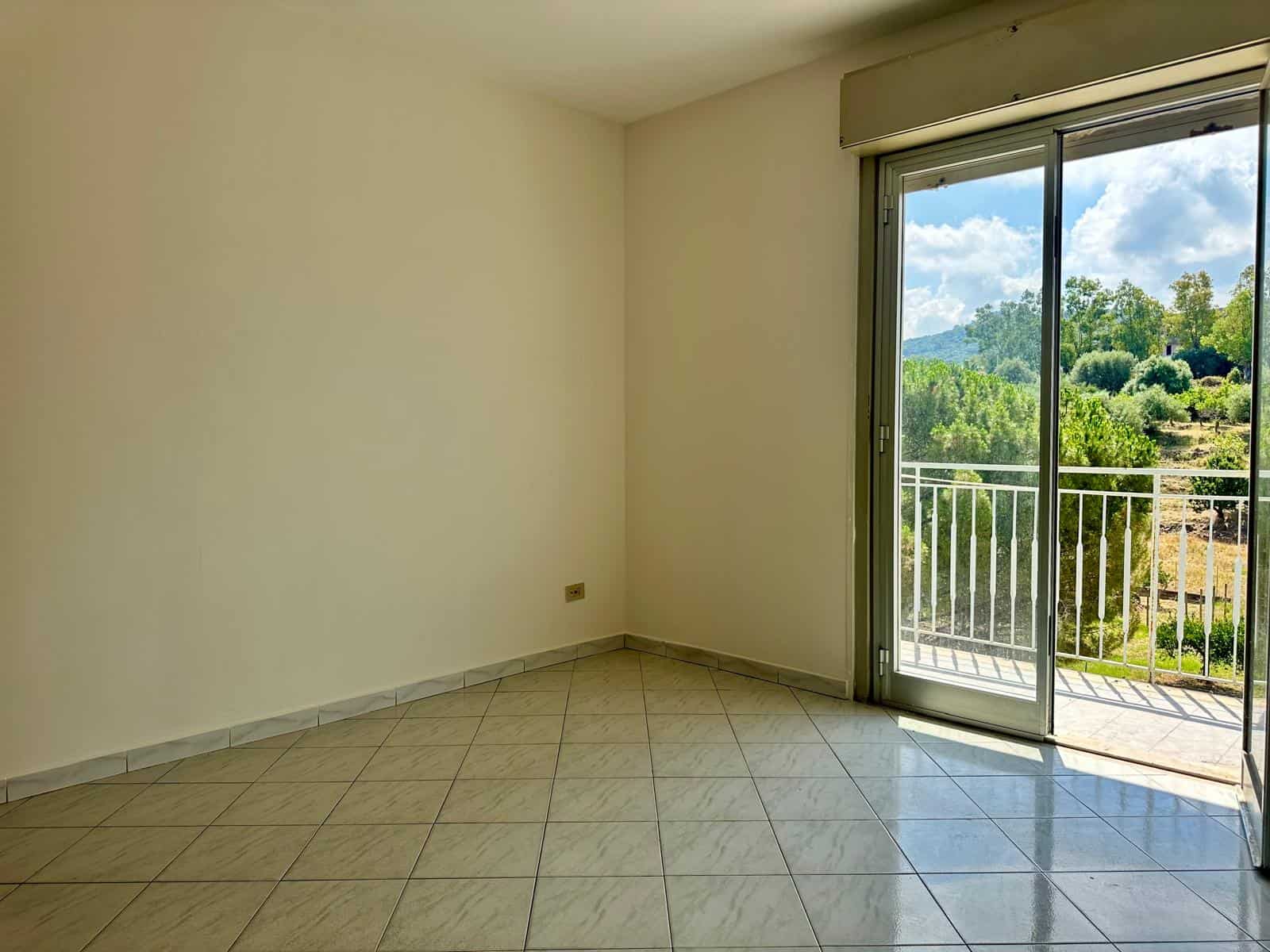 Stanza luminosa con grande porta finestra che dà su un balcone con vista su natura e colline.