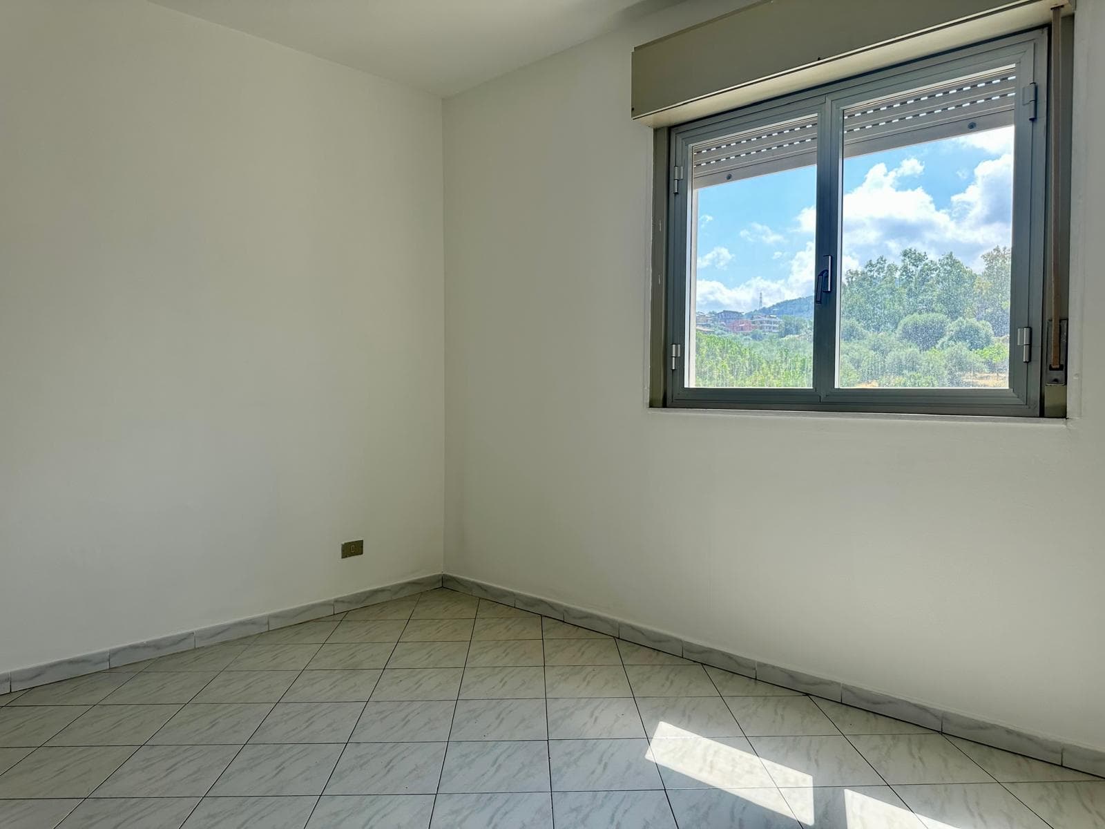 Stanza luminosa con grande finestra che mostra un paesaggio verde e cielo azzurro, ideale per appartamento in vendita in via Delle Ginestre.