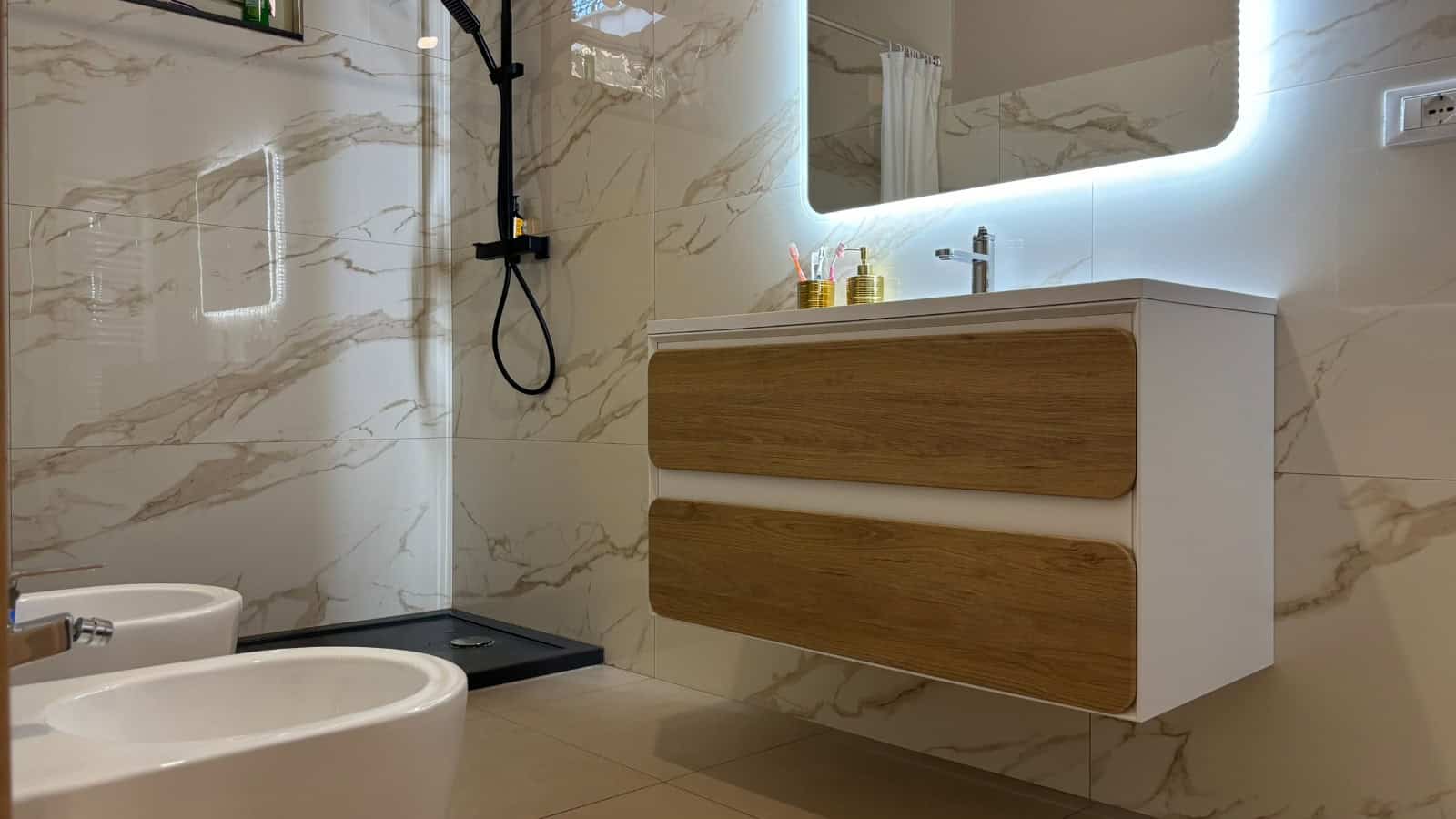 Bagno moderno con lavabo sospeso, specchio con illuminazione a LED e pareti in marmo chiaro.