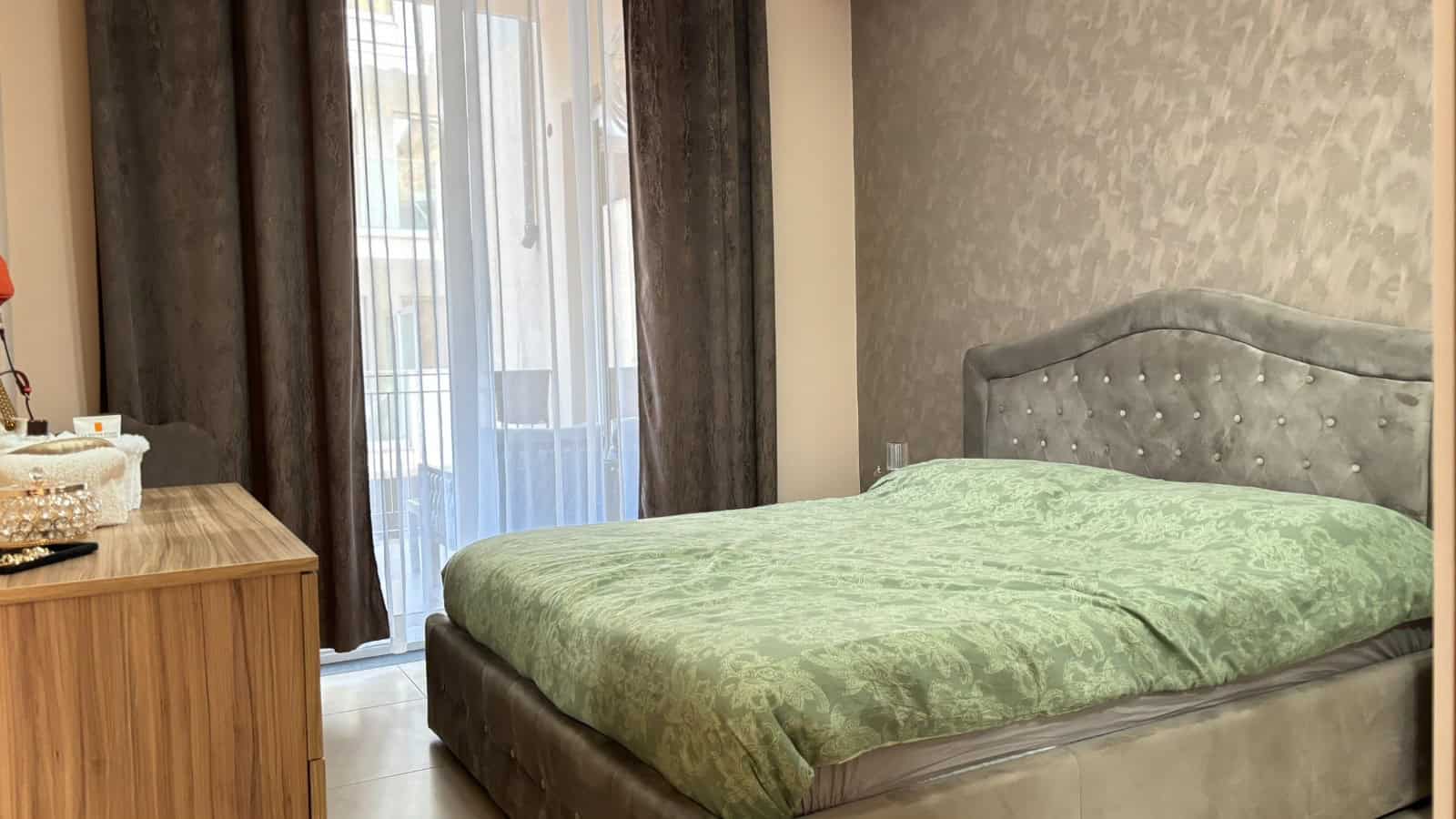 Camera da letto moderna con letto imbottito e finestra con tende scure e leggere, vista su balcone.