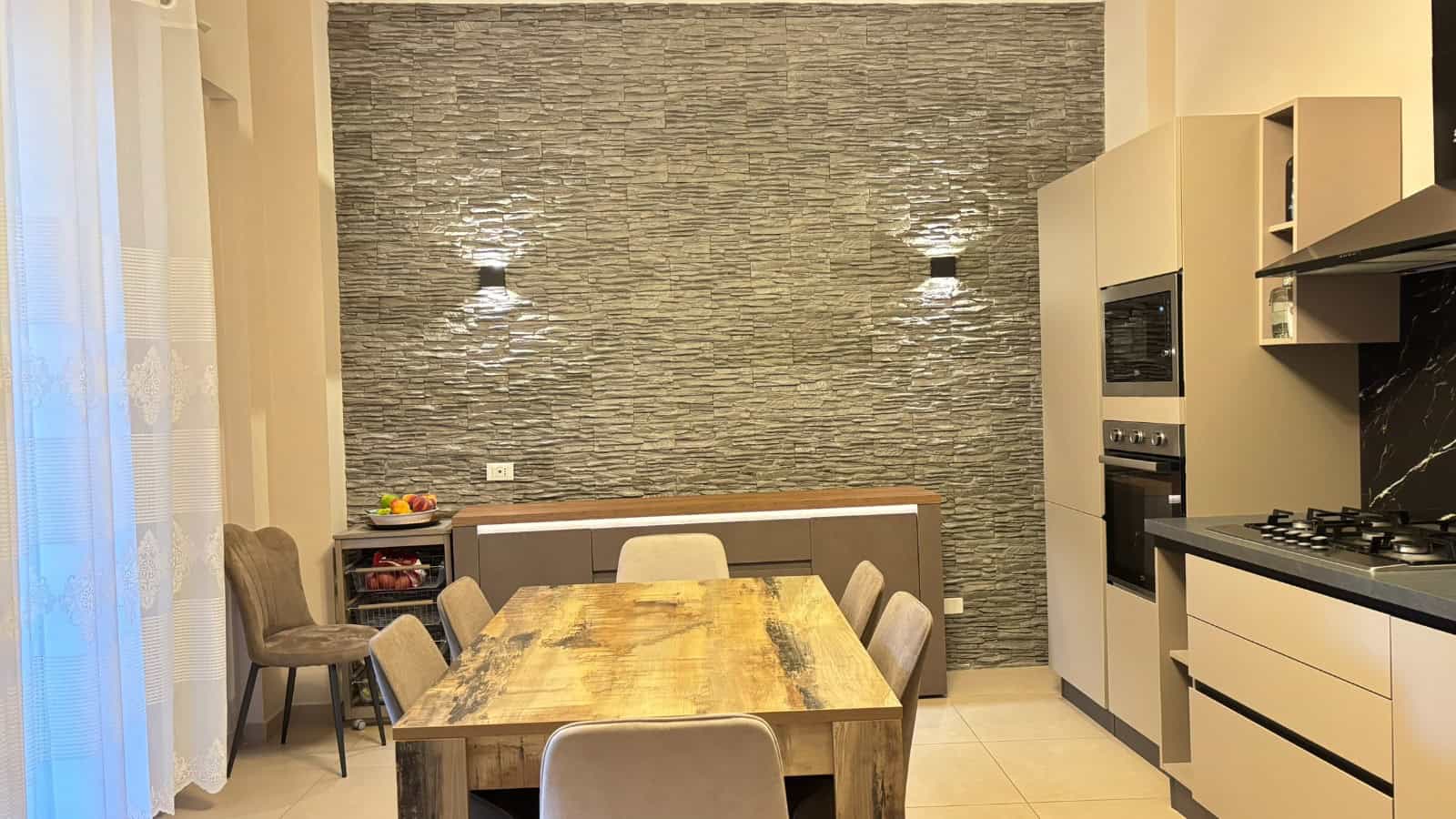Interno moderno di un appartamento in vendita a Milazzo, cucina con parete in pietra e zona pranzo con tavolo in legno e sedie.