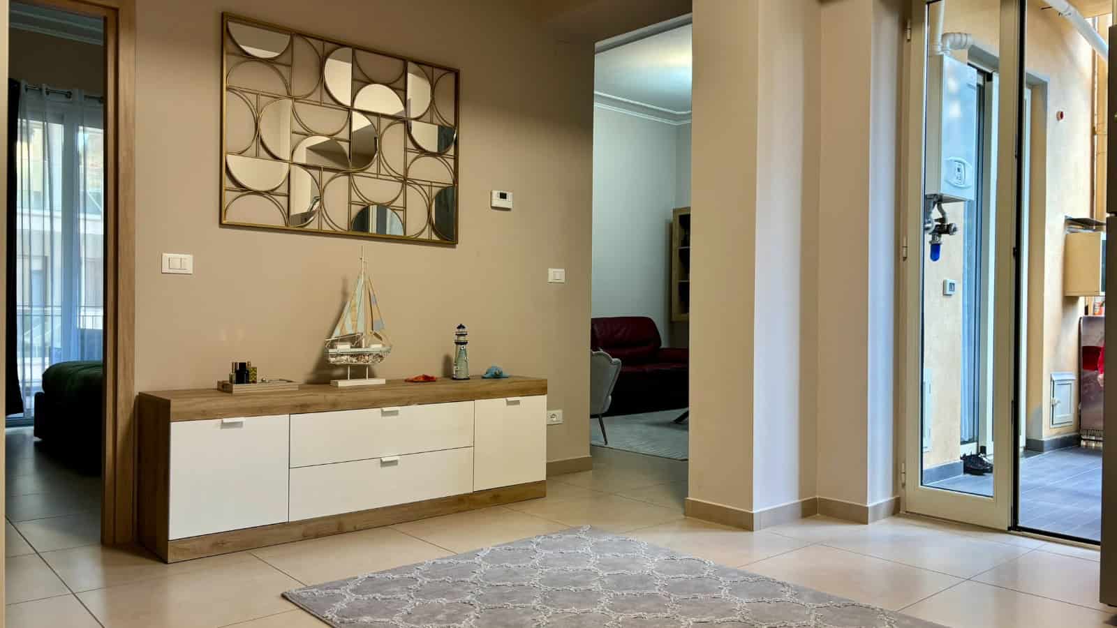 Interno di un appartamento in vendita a Milazzo, con parete decorativa e mobile basso, ingresso luminoso e accesso a un balcone.