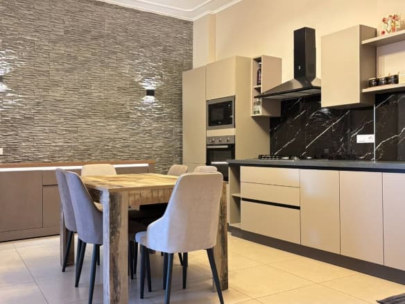 Cucina moderna con mobili beige e parete in pietra, tavolo in legno e sedie imbottite in tessuto grigio, ideale per appartamento in vendita a Milazzo.