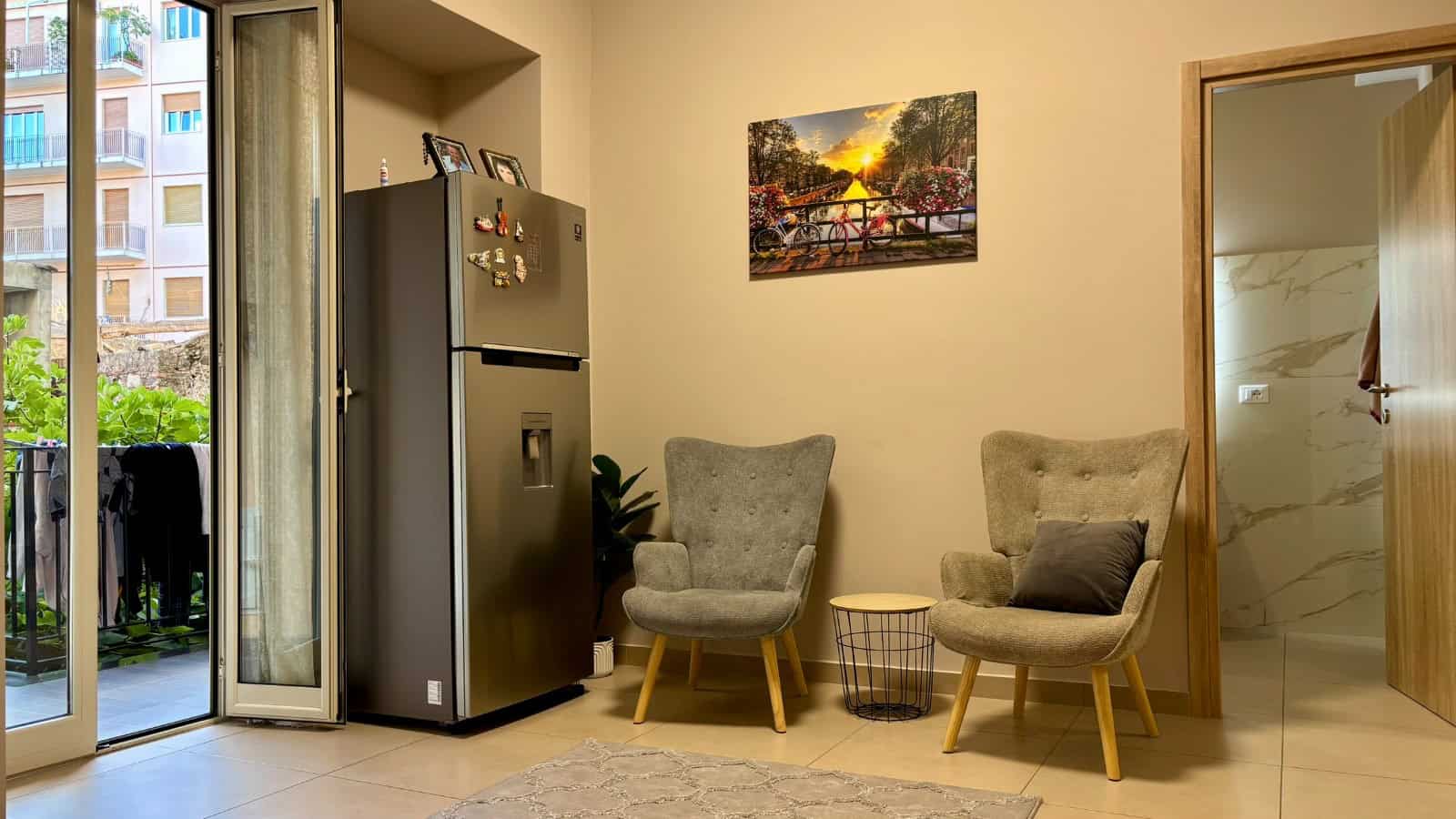 Interno di un appartamento con due poltrone grigie, frigorifero e vista su balcone con piante e edifici.
