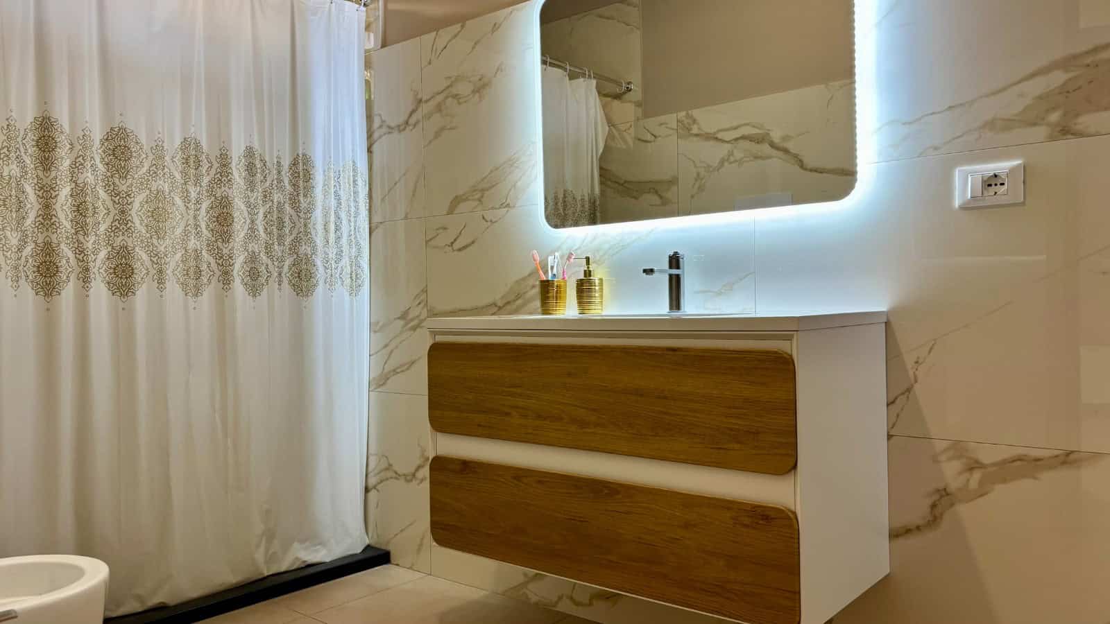 Bagno moderno con lavabo sospeso, specchio con illuminazione a LED e pareti in marmo chiaro.