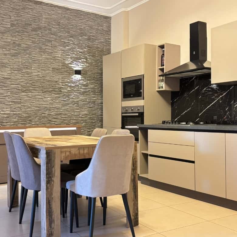 Cucina moderna con mobili beige e parete in pietra, tavolo in legno e sedie imbottite in tessuto grigio, ideale per appartamento in vendita a Milazzo.