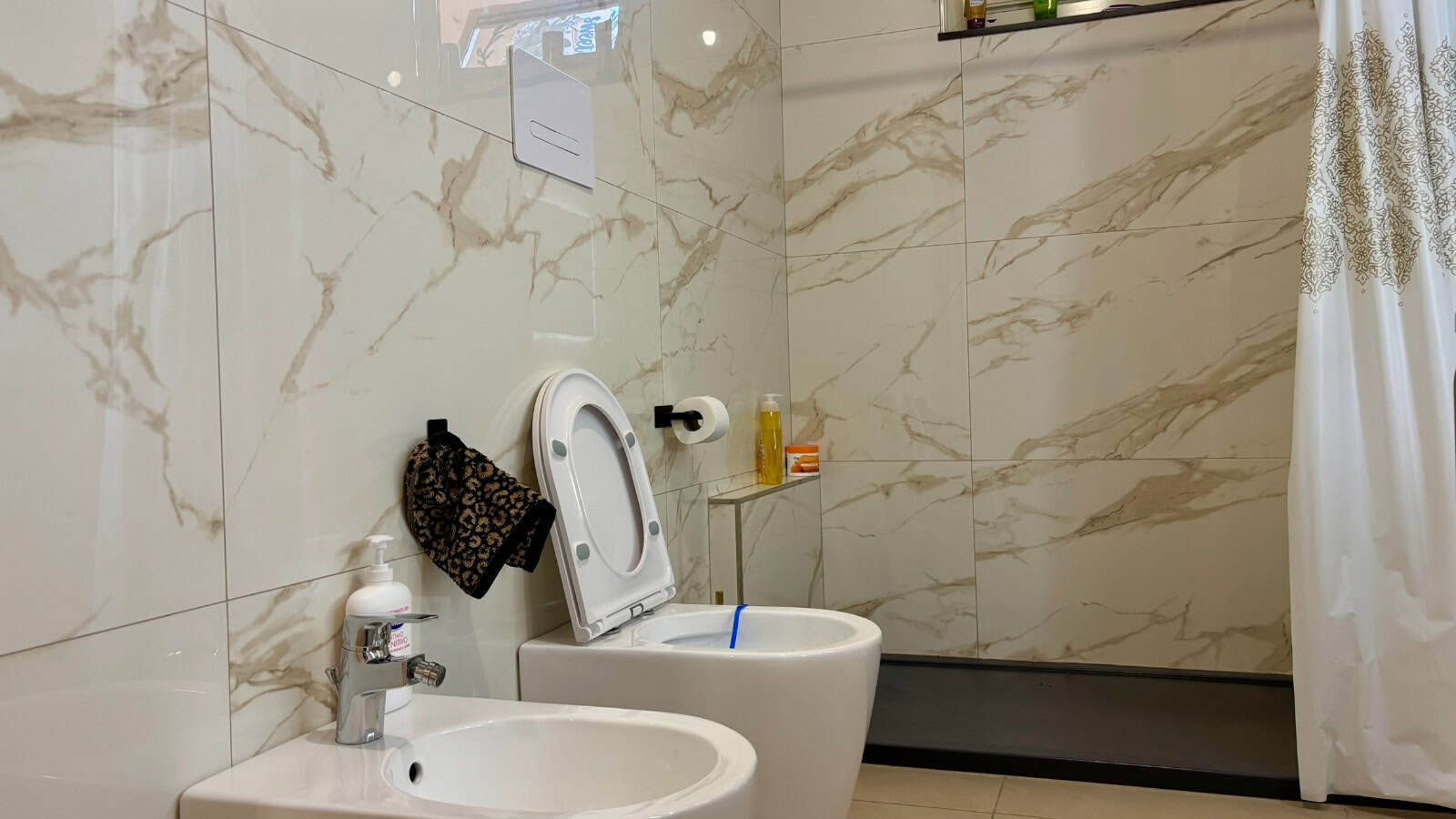 Bagno moderno con pareti in marmo e sanitari bianchi, dotato di doccia e accessori essenziali.