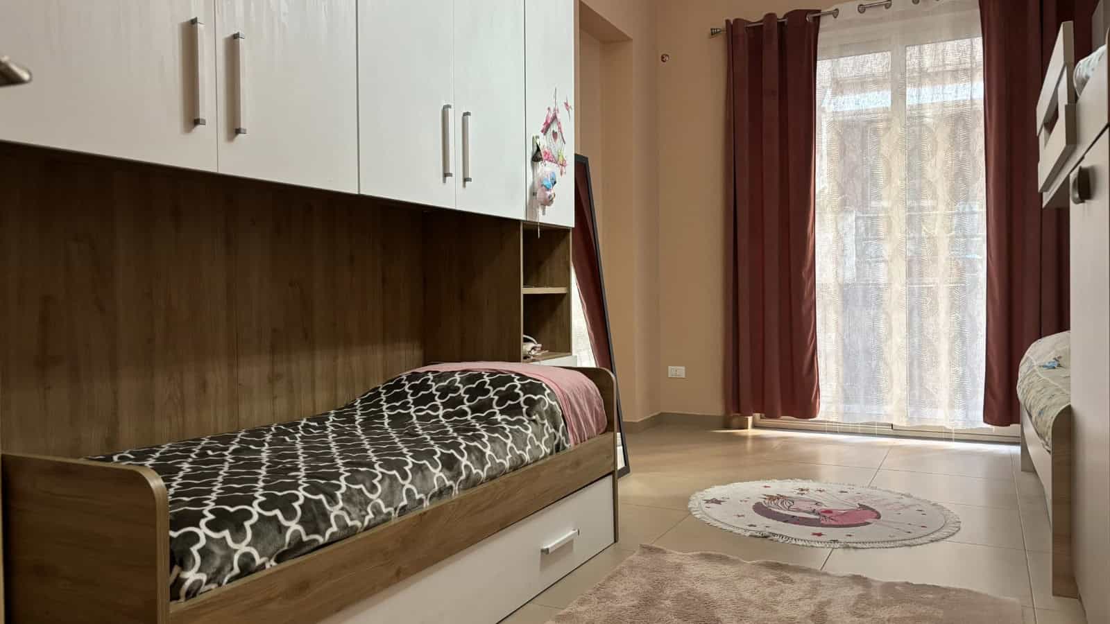 Interno di un appartamento con letto a castello, armadi e grande finestra con tende rosse.