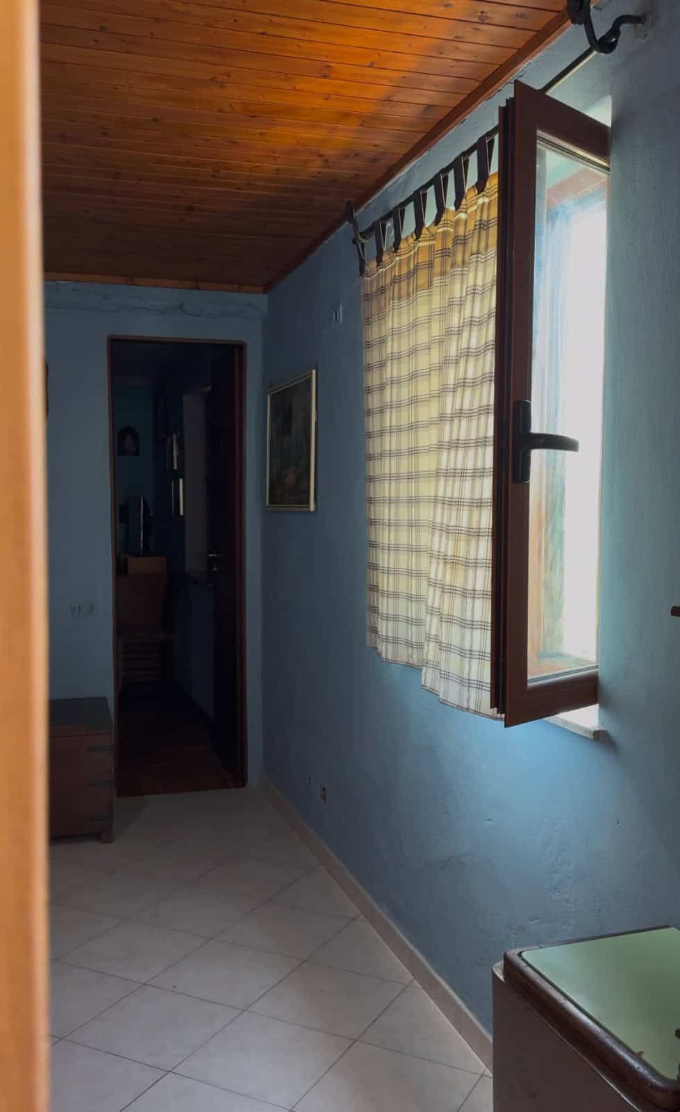 Interno di una casa a Quattropani, Lipari, con pareti azzurre, finestra con tende a quadretti e soffitto in legno.