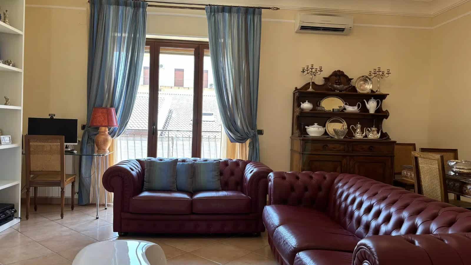 Elegante soggiorno con divani in pelle rosso bordeaux, grandi finestre con tende azzurre e arredi classici in stile tradizionale.