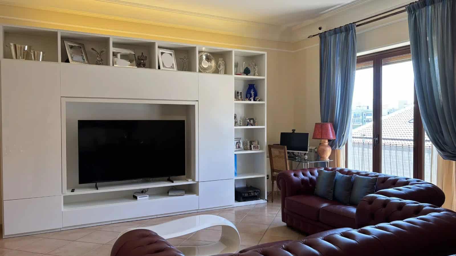 Soggiorno luminoso con divani rossi, TV e libreria bianca, con grandi finestre e tende blu, in un appartamento in vendita a Milazzo.