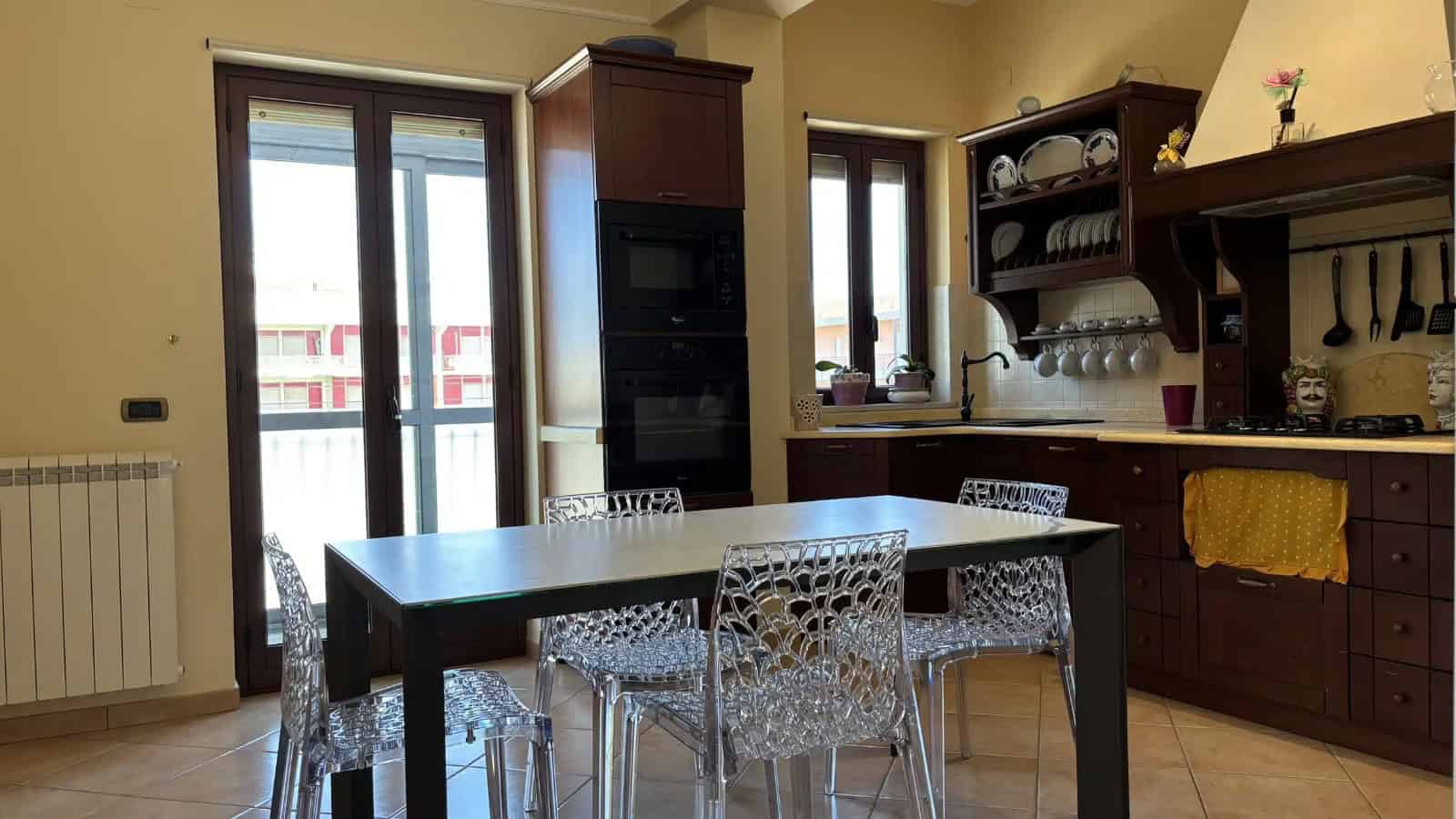 Interno di un appartamento con cucina e zona pranzo, ampie finestre e mobili in legno.