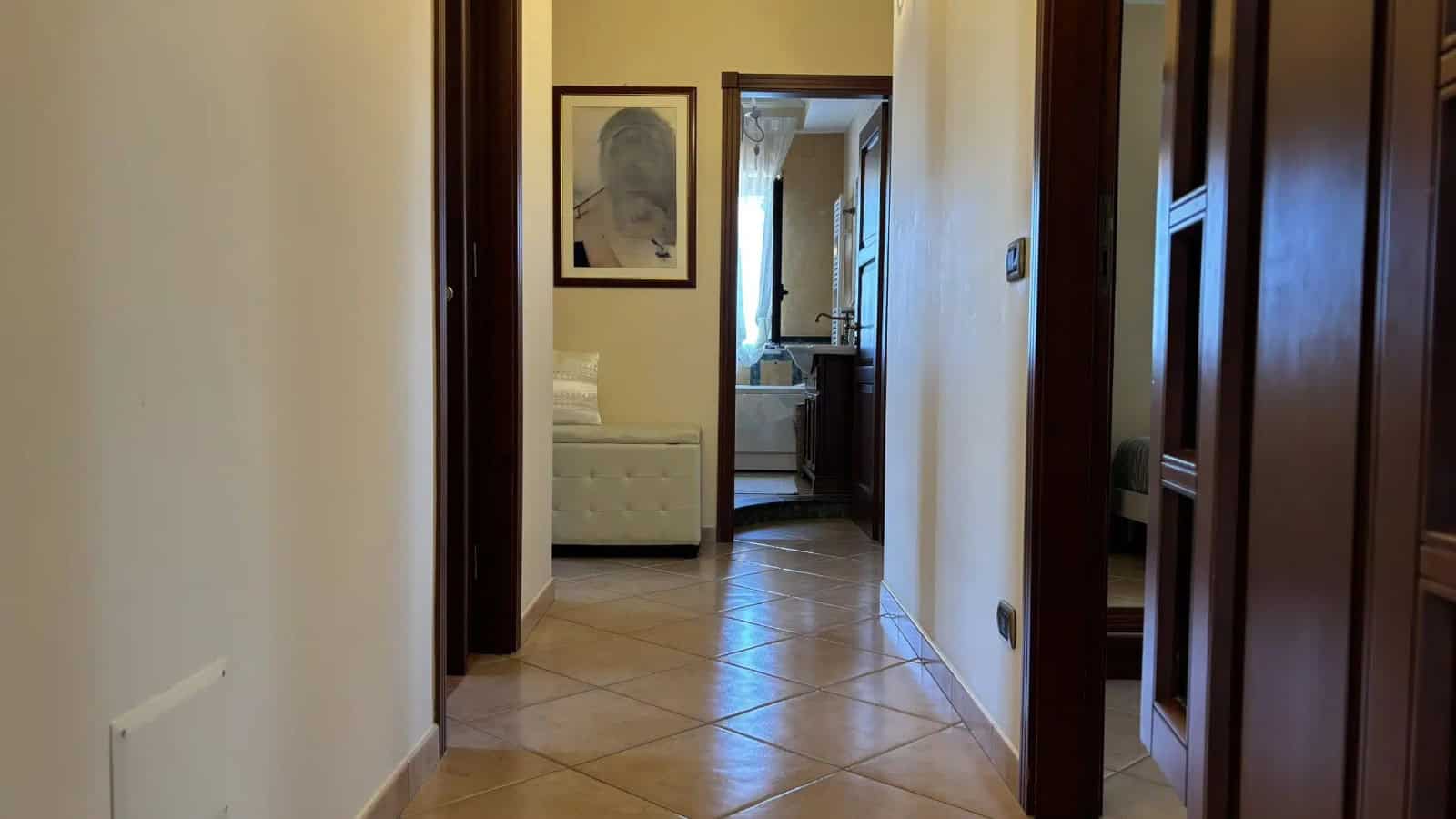 Ingresso di un appartamento con pareti beige, pavimento in piastrelle e porte in legno scuro, vista verso la zona giorno e una finestra luminosa.