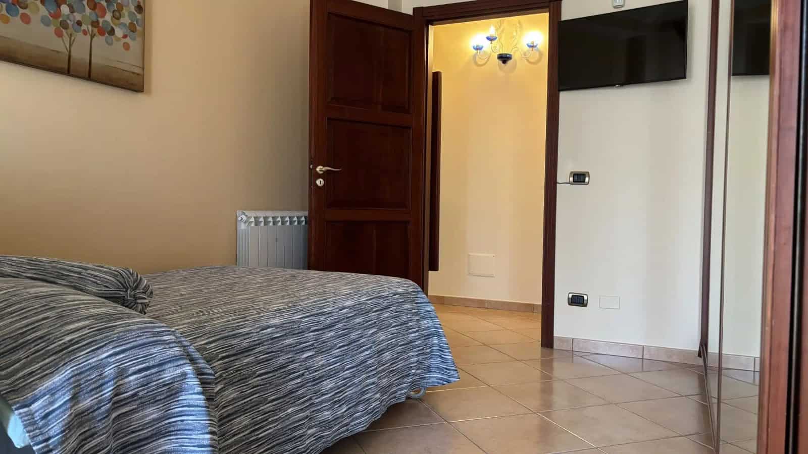 Camera che mostra una camera da letto con letto matrimoniale, pareti beige e porta in legno aperta verso un ingresso.