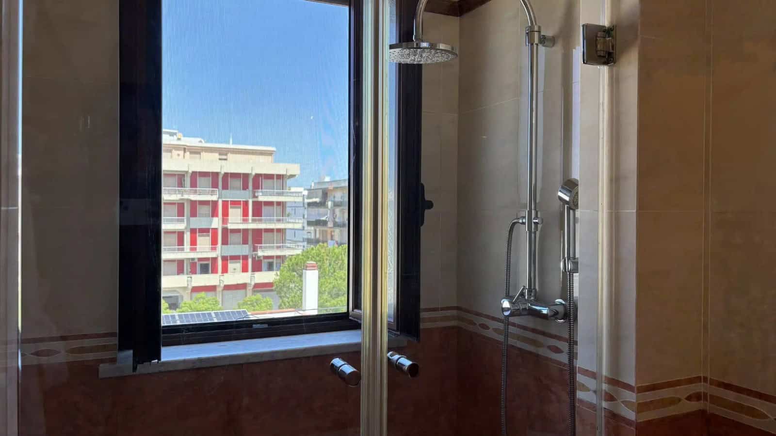 Vista dalla finestra di un bagno con doccia, edifici e cielo azzurro all'esterno.