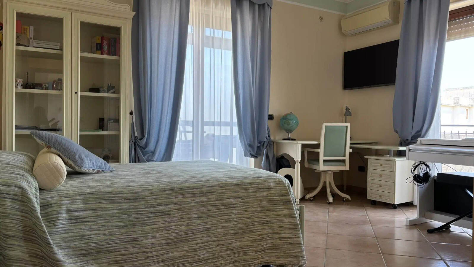 Interno di un appartamento con camera da letto luminosa, arredi moderni e vista su balcone con tende azzurre.