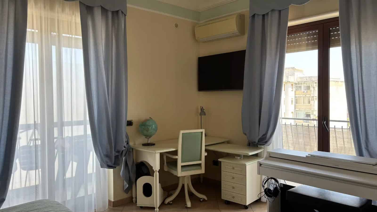 Interno di un appartamento con scrivania, sedia, televisore e balcone con tende azzurre.