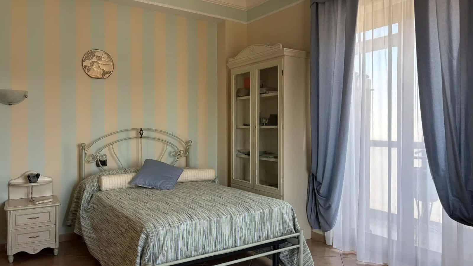 Camera da letto luminosa con pareti a strisce azzurre e beige, letto matrimoniale con cuscino blu, armadio in vetro e tende blu, vista su balcone.