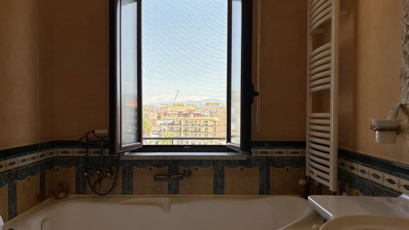 Vista dalla finestra di un bagno con vasca, con panorama urbano di Milazzo sullo sfondo.