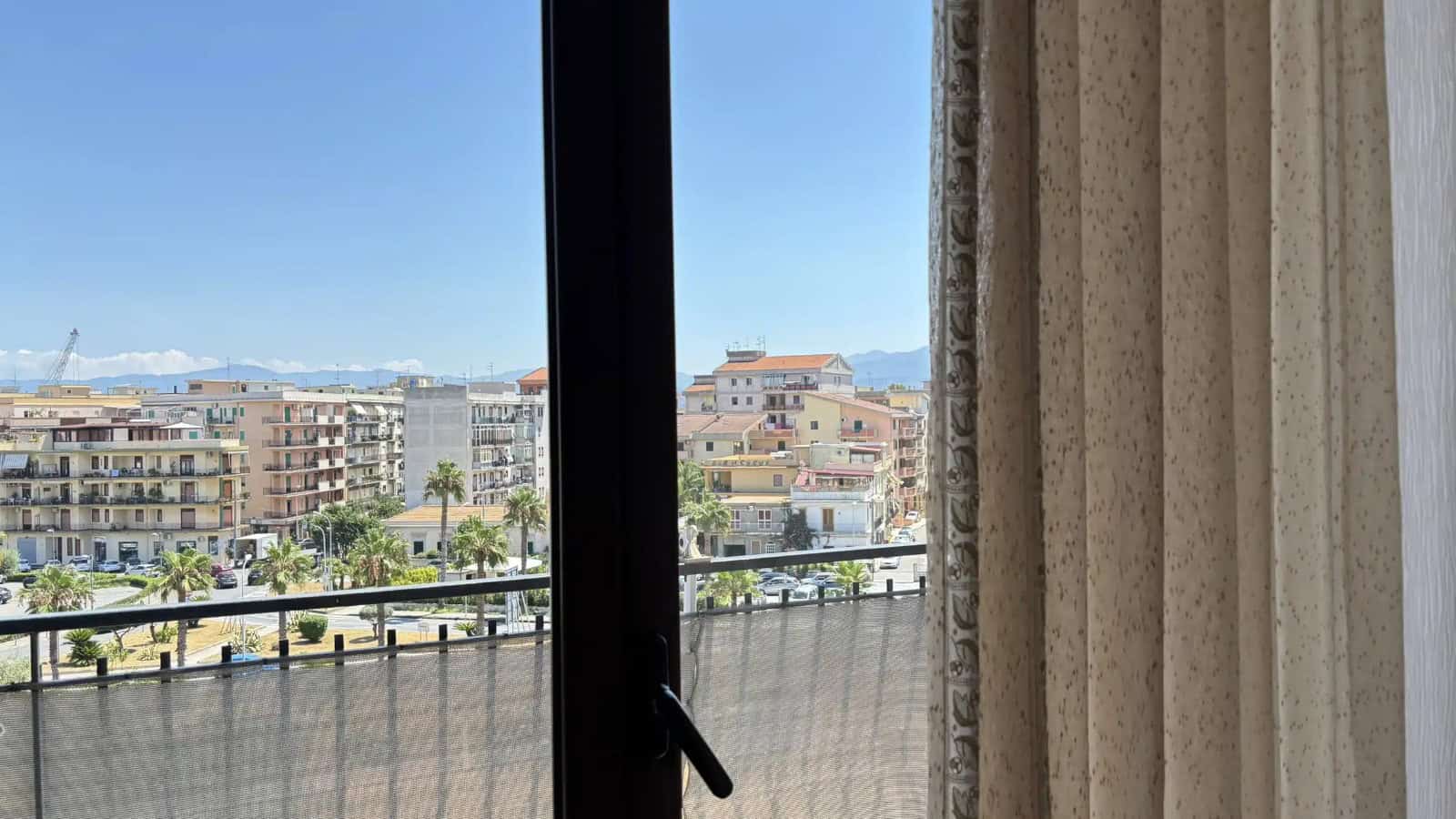 Vista panoramica di Milazzo vista da una finestra con tende beige, con edifici e mare sullo sfondo.