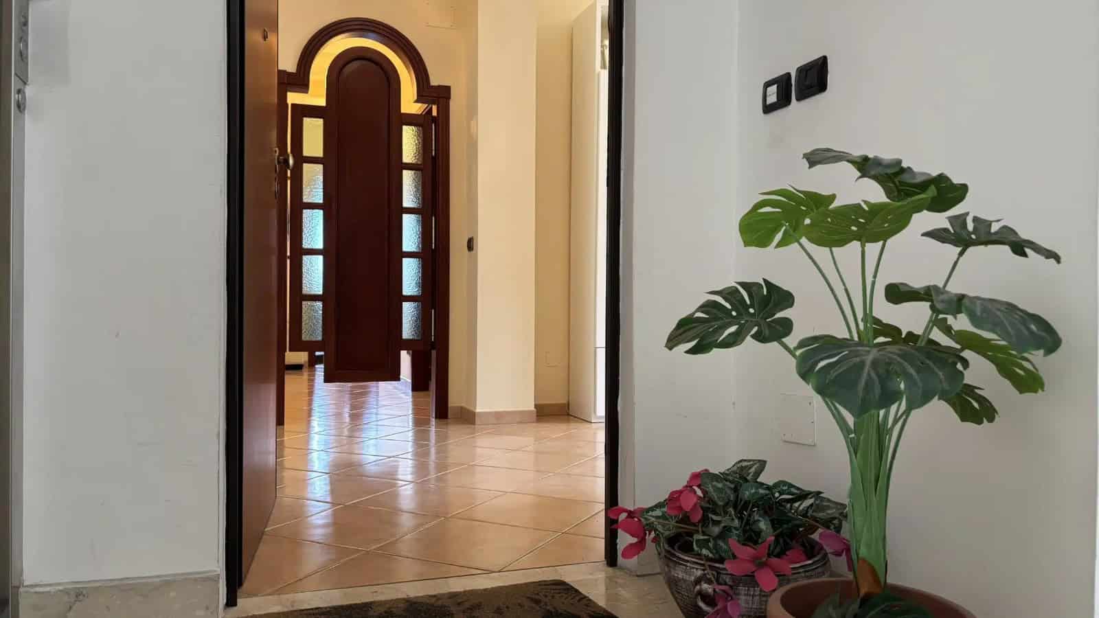 Ingresso di un appartamento con porta in legno e finestra decorativa, arredato con piante verdi e fiori.