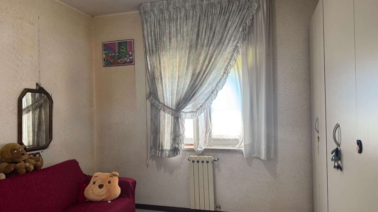 Interno di un appartamento con finestra, tende leggere e armadio bianco, decorato con un quadro e un divano con cuscino a forma di orsetto.