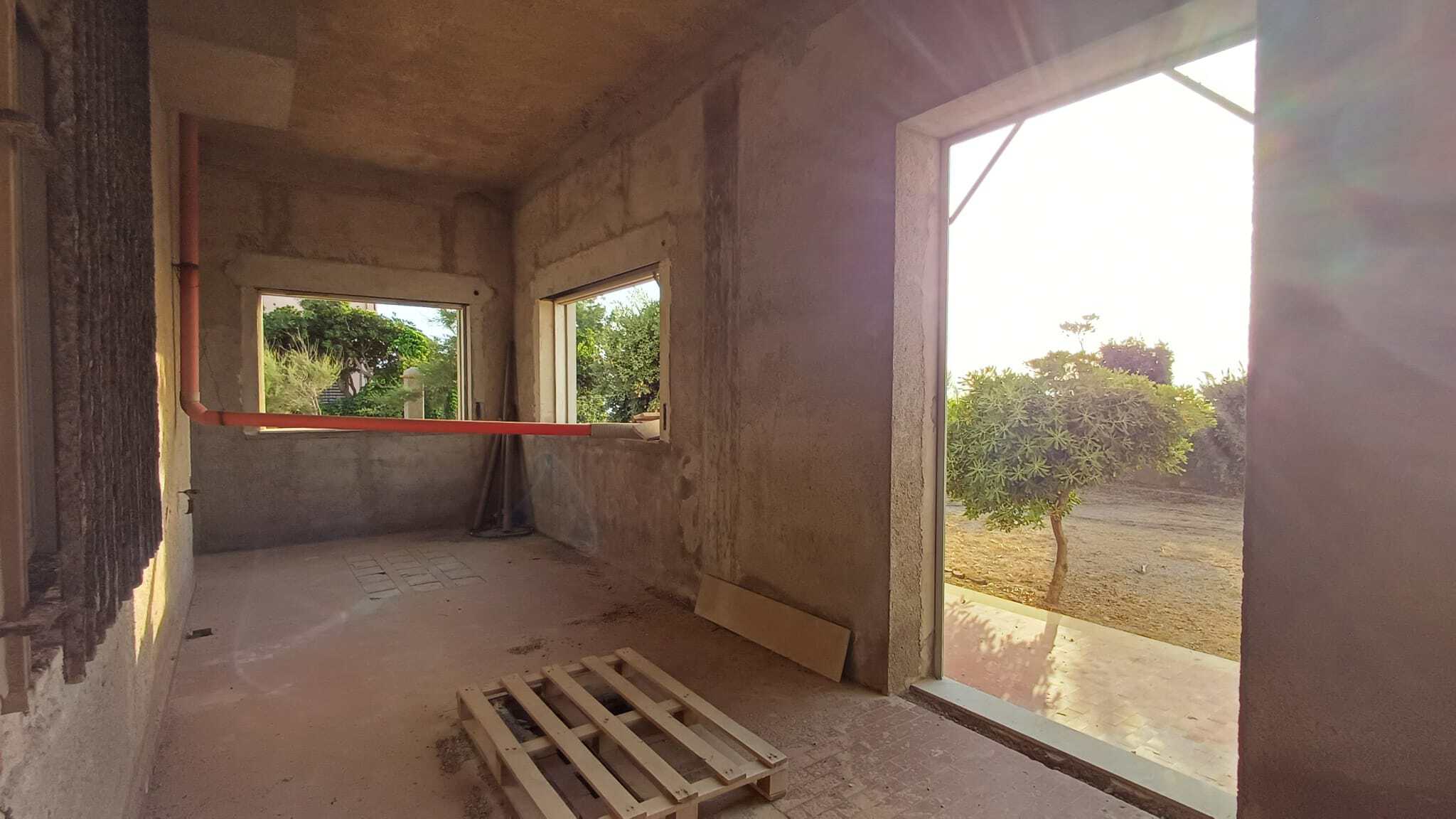 Interno di un locale commerciale in costruzione con grandi finestre e vista su un giardino con alberi.